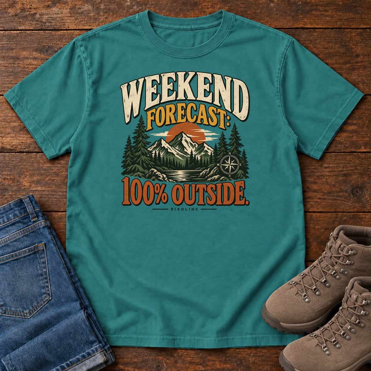 Emerald Weekend Forecast 100 Percent Outside T-Shirt with mountains, evergreens, an orange sun and a compass beneath bold lettering; front flat lay on wood.