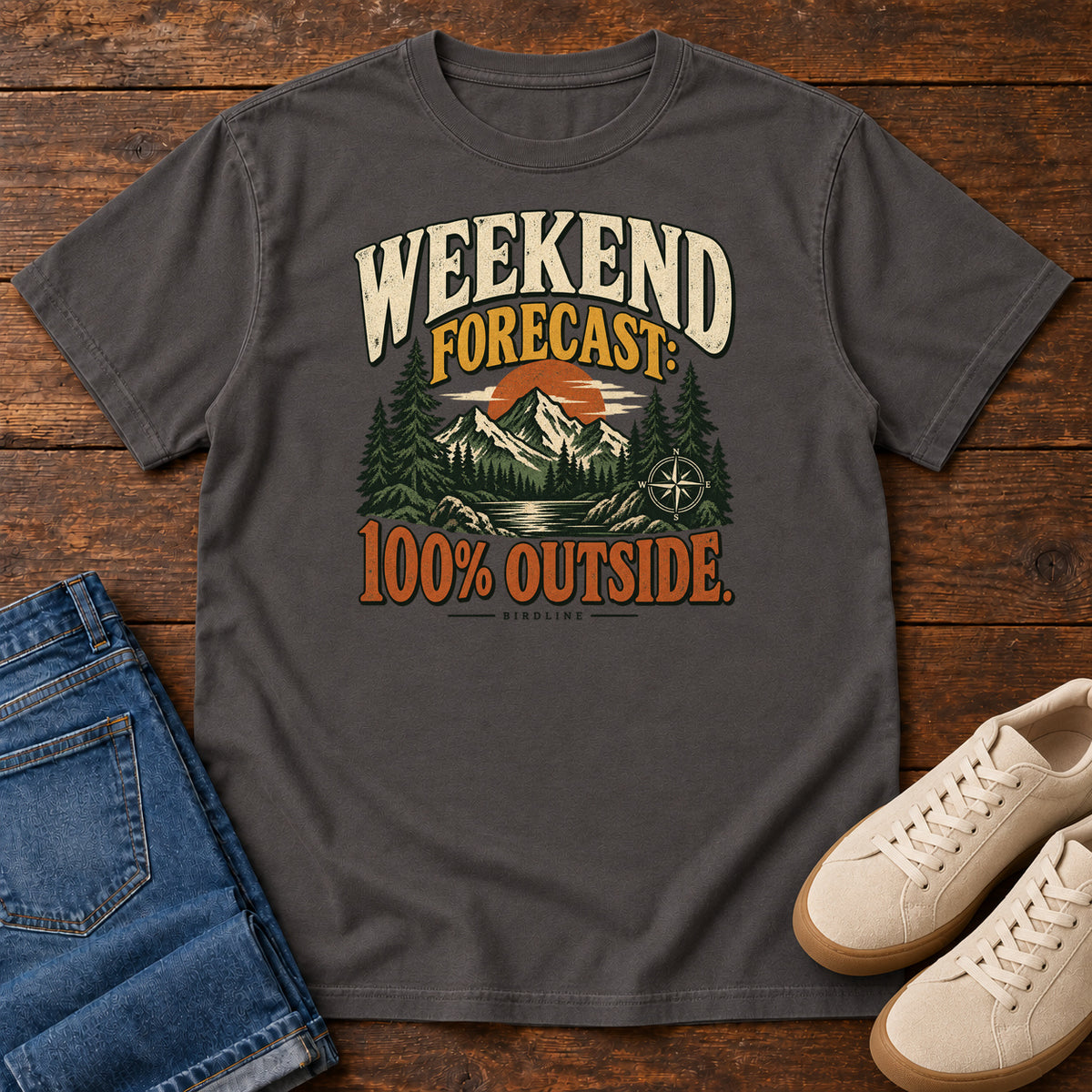 Graphite Weekend Forecast 100 Percent Outside T-Shirt with mountains, evergreens, an orange sun and a compass beneath bold lettering; front flat lay on wood.