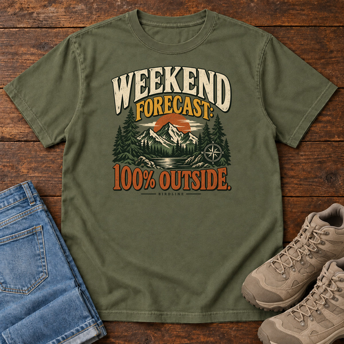 Hemp Weekend Forecast 100 Percent Outside T-Shirt with mountains, evergreens, an orange sun and a compass beneath bold lettering; front flat lay on wood.