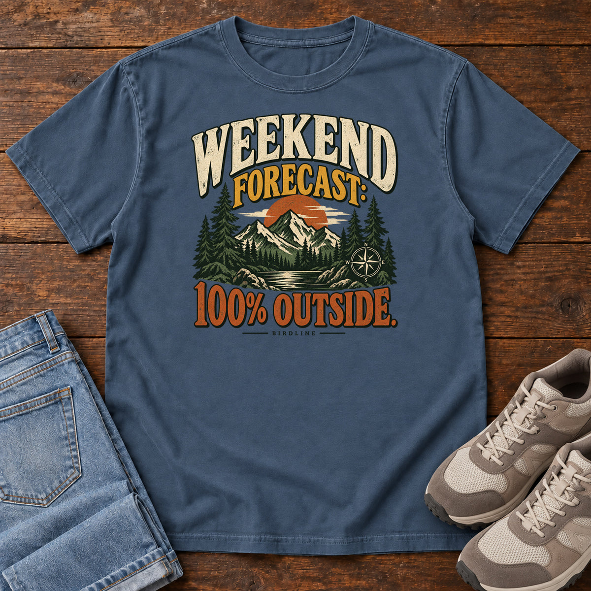 Midnight Weekend Forecast 100 Percent Outside T-Shirt with mountains, evergreens, an orange sun and a compass beneath bold lettering; front flat lay on wood.