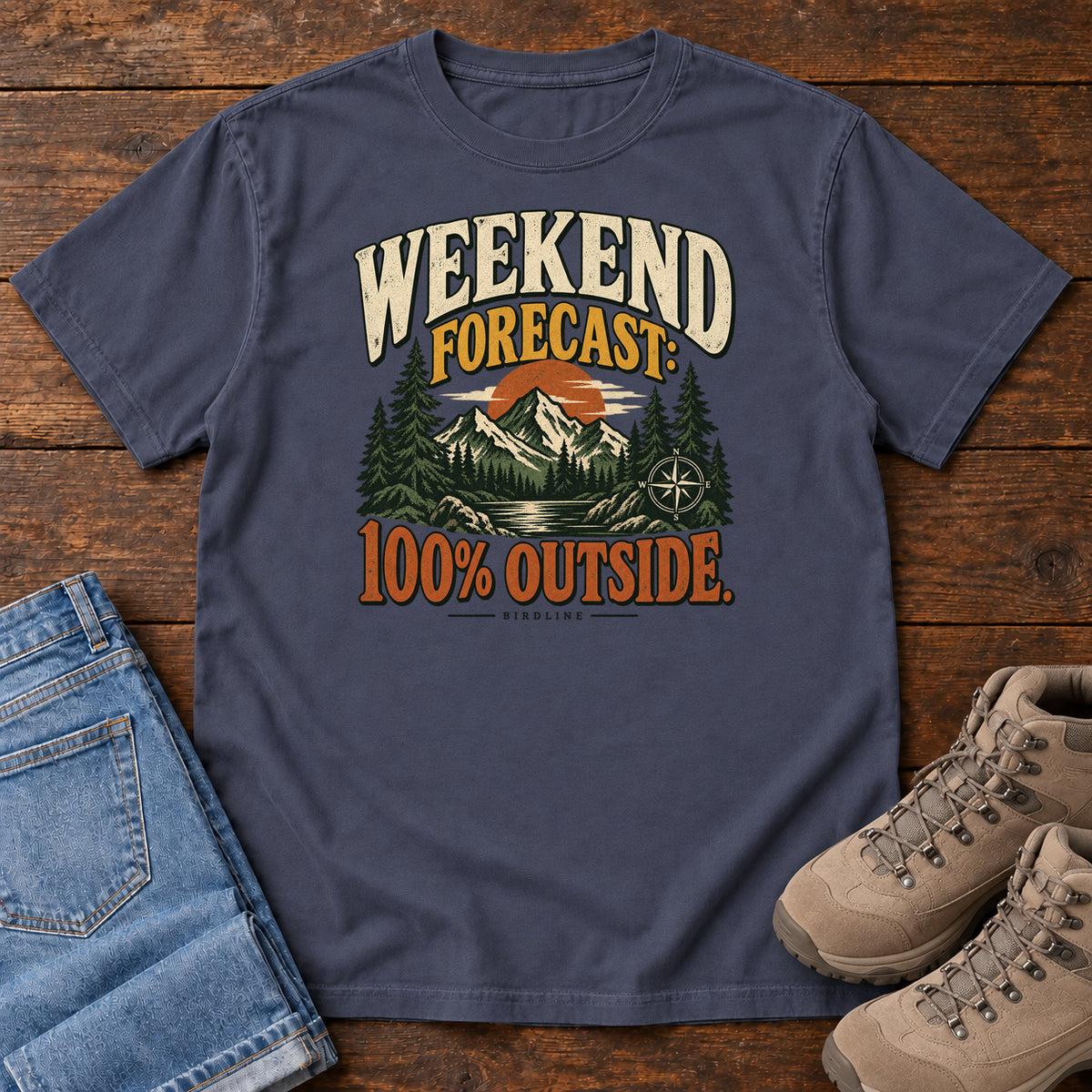 Navy Weekend Forecast 100 Percent Outside T-Shirt with mountains, evergreens, an orange sun and a compass beneath bold lettering; front flat lay on wood.