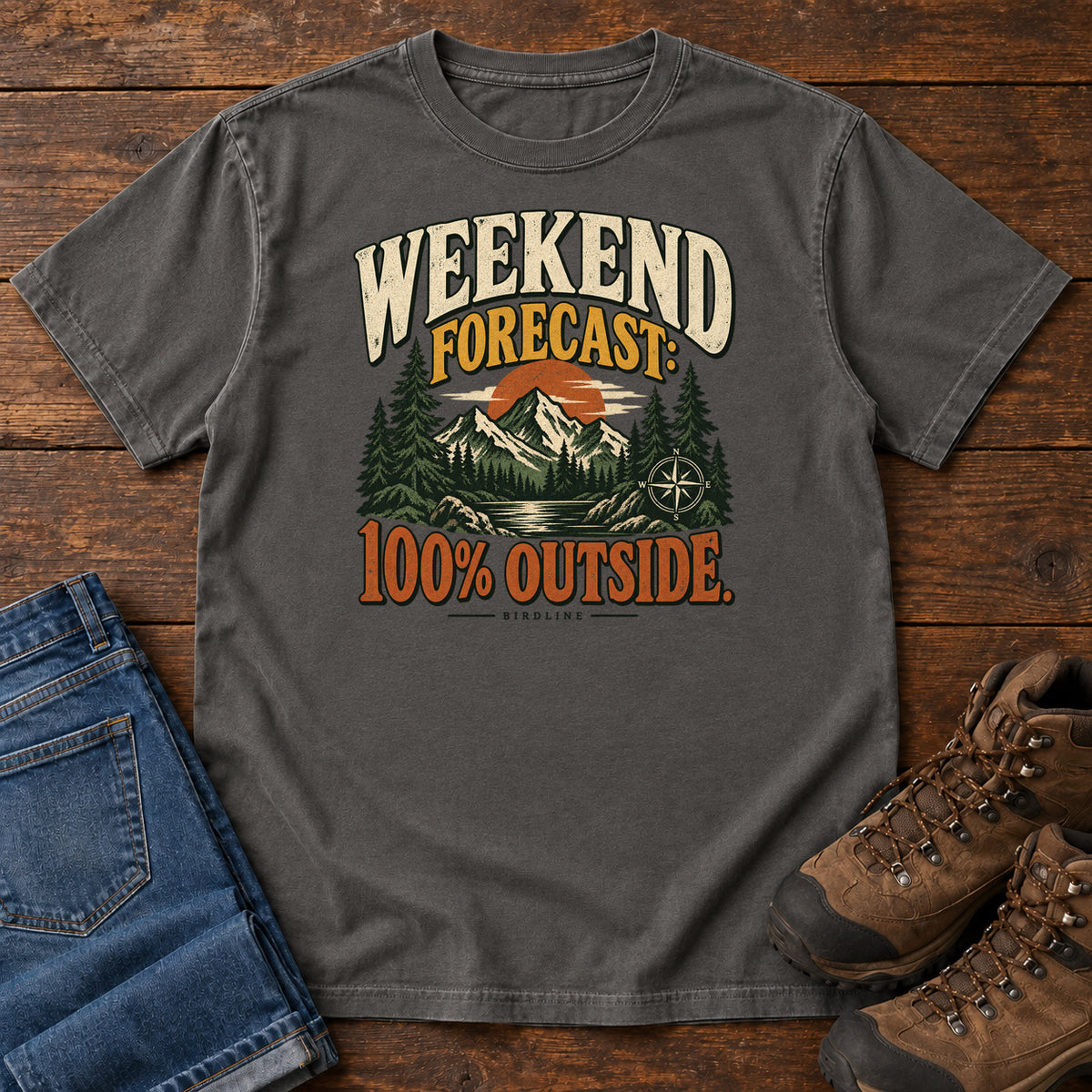 Pepper Weekend Forecast 100 Percent Outside T-Shirt with mountains, evergreens, an orange sun and a compass beneath bold lettering; front flat lay on wood.