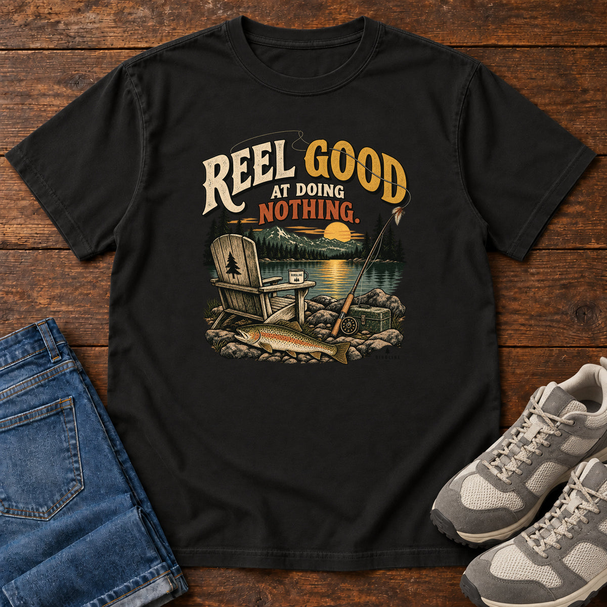 Black Reel Good at Doing Nothing T-Shirt with a wooden chair and fishing rod beside a lake at sunset, front flat lay on wood.