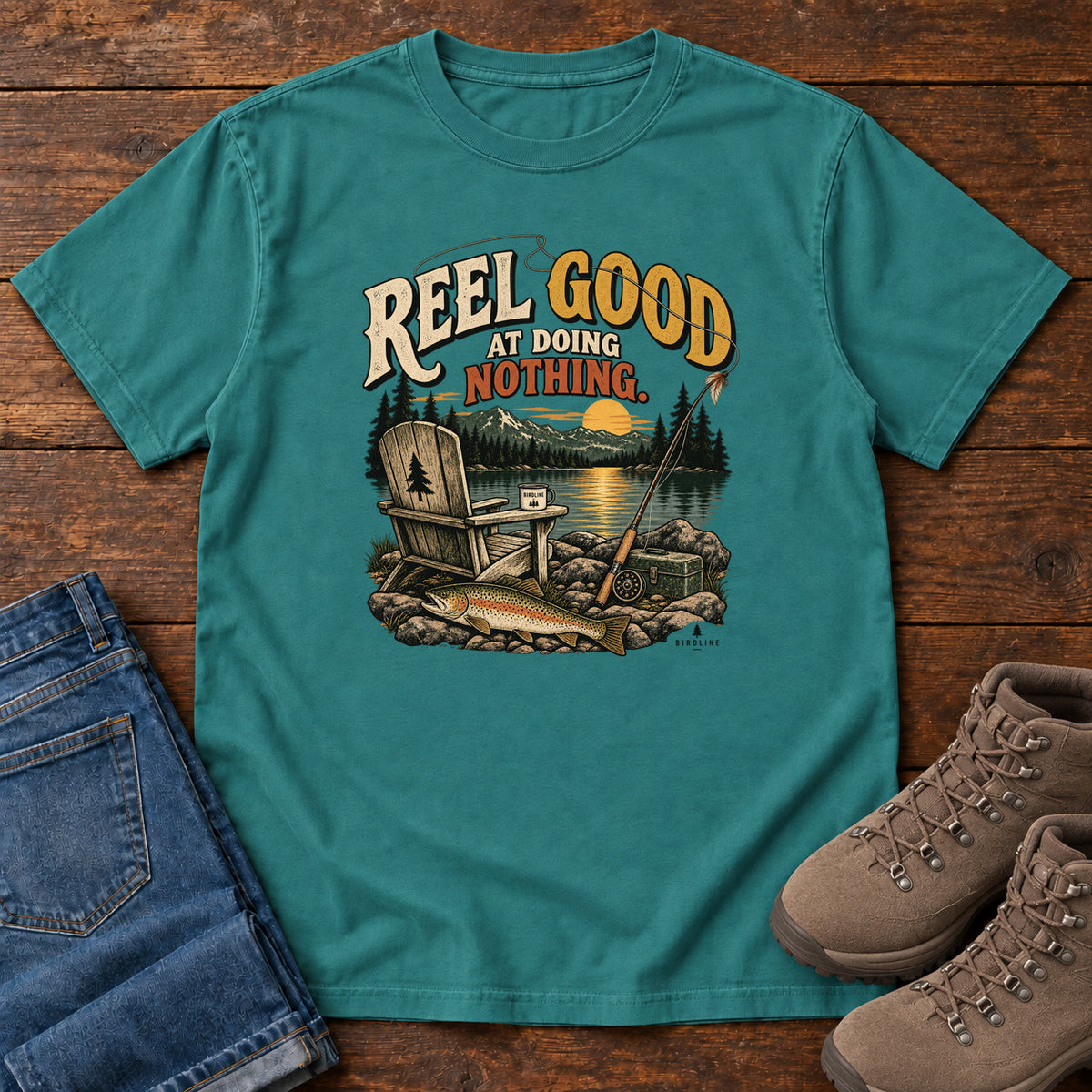 Emerald Reel Good at Doing Nothing T-Shirt with a wooden chair and fishing rod beside a lake at sunset, front flat lay on wood.