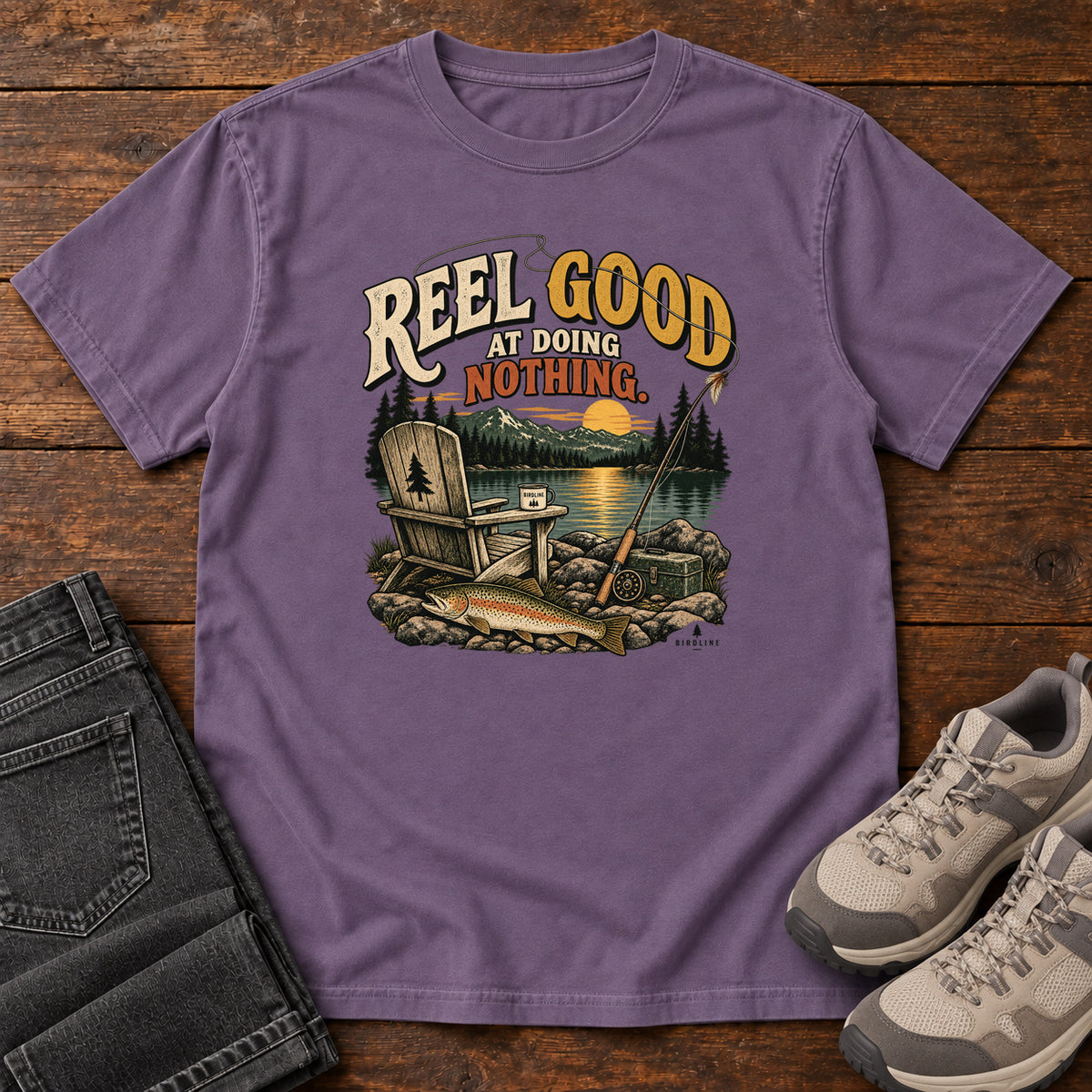 Grape Reel Good at Doing Nothing T-Shirt with a wooden chair and fishing rod beside a lake at sunset, front flat lay on wood.