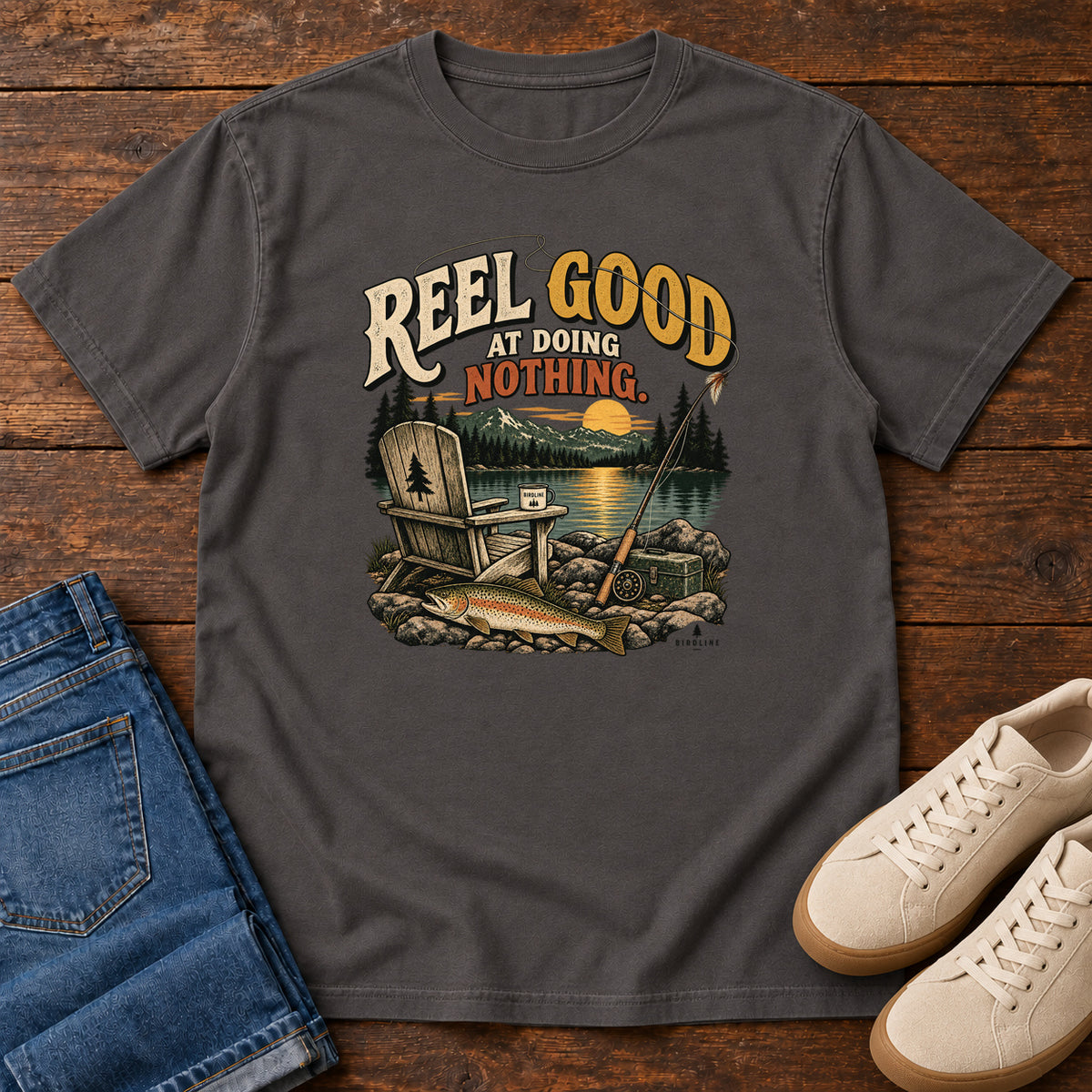 Graphite Reel Good at Doing Nothing T-Shirt with a wooden chair and fishing rod beside a lake at sunset, front flat lay on wood.