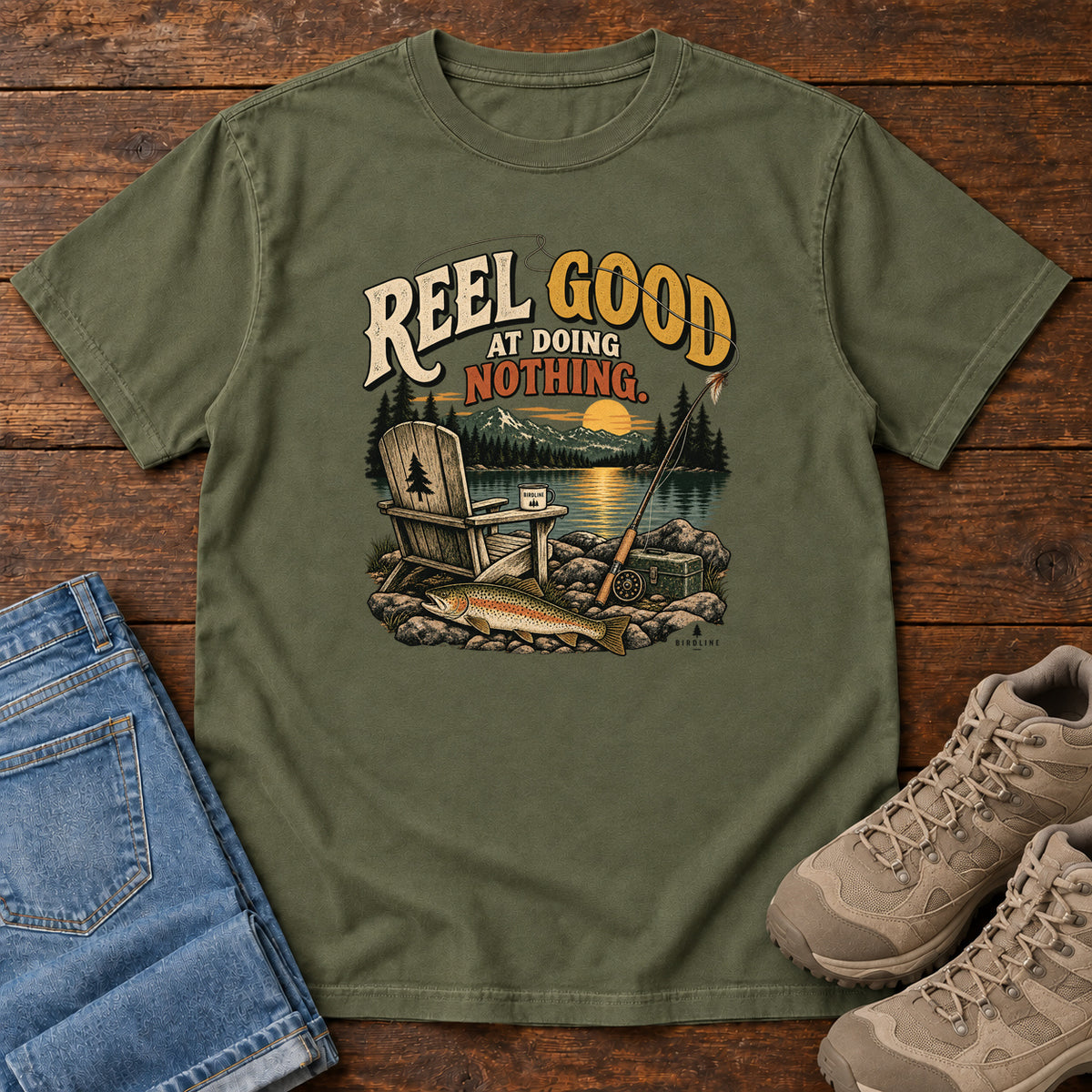 Hemp Reel Good at Doing Nothing T-Shirt with a wooden chair and fishing rod beside a lake at sunset, front flat lay on wood.