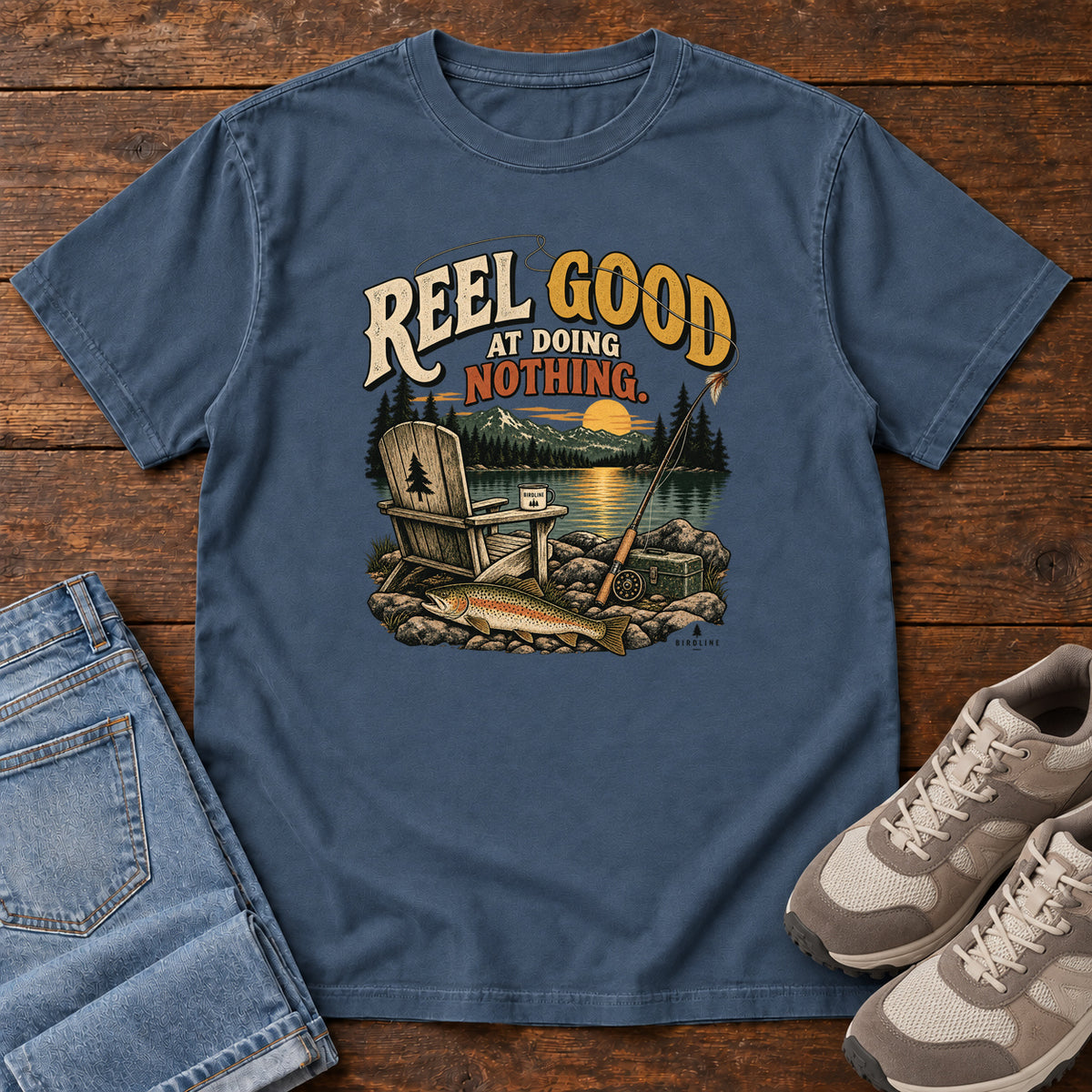 Midnight Reel Good at Doing Nothing T-Shirt with a wooden chair and fishing rod beside a lake at sunset, front flat lay on wood.