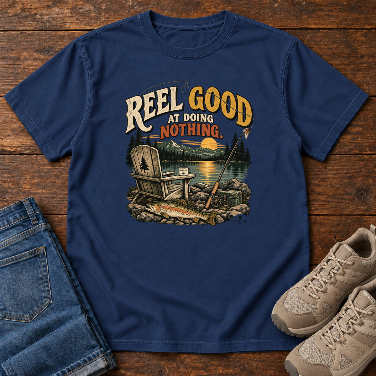 True Navy Reel Good at Doing Nothing T-Shirt with a wooden chair and fishing rod beside a lake at sunset, front flat lay on wood.