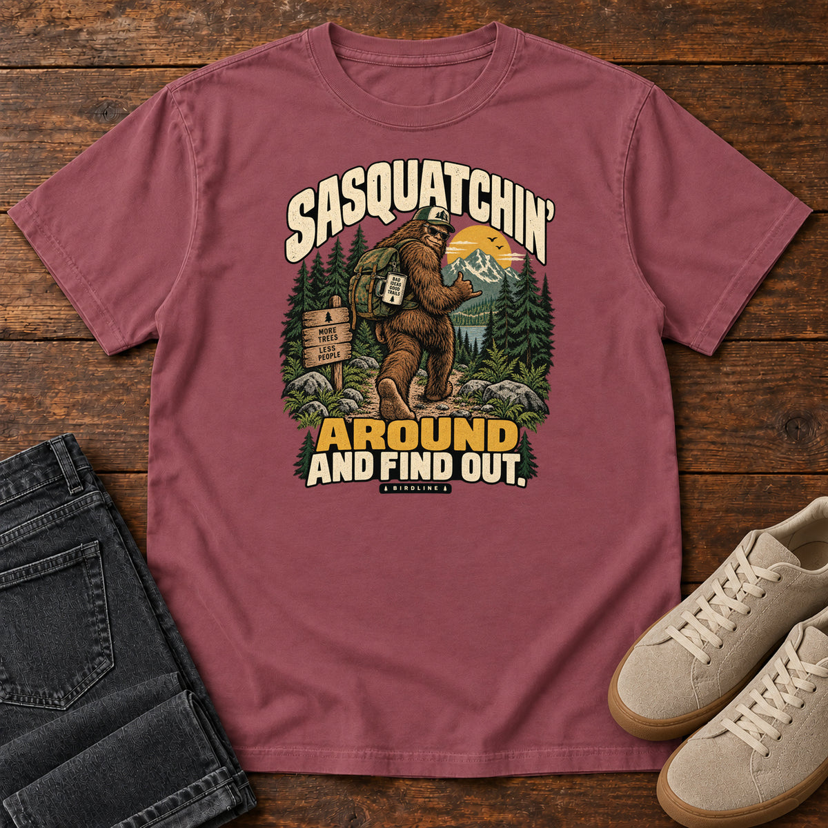 Brick Sasquatchin Around and Find Out T-Shirt with a backpack-wearing Sasquatch on a forest trail with cream-and-gold lettering, front flat lay on wood.