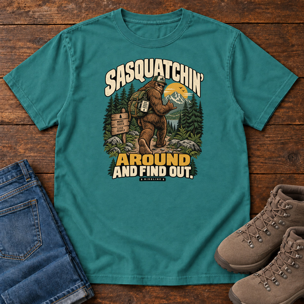 Emerald Sasquatchin Around and Find Out T-Shirt with a backpack-wearing Sasquatch on a forest trail with cream-and-gold lettering, front flat lay on wood.