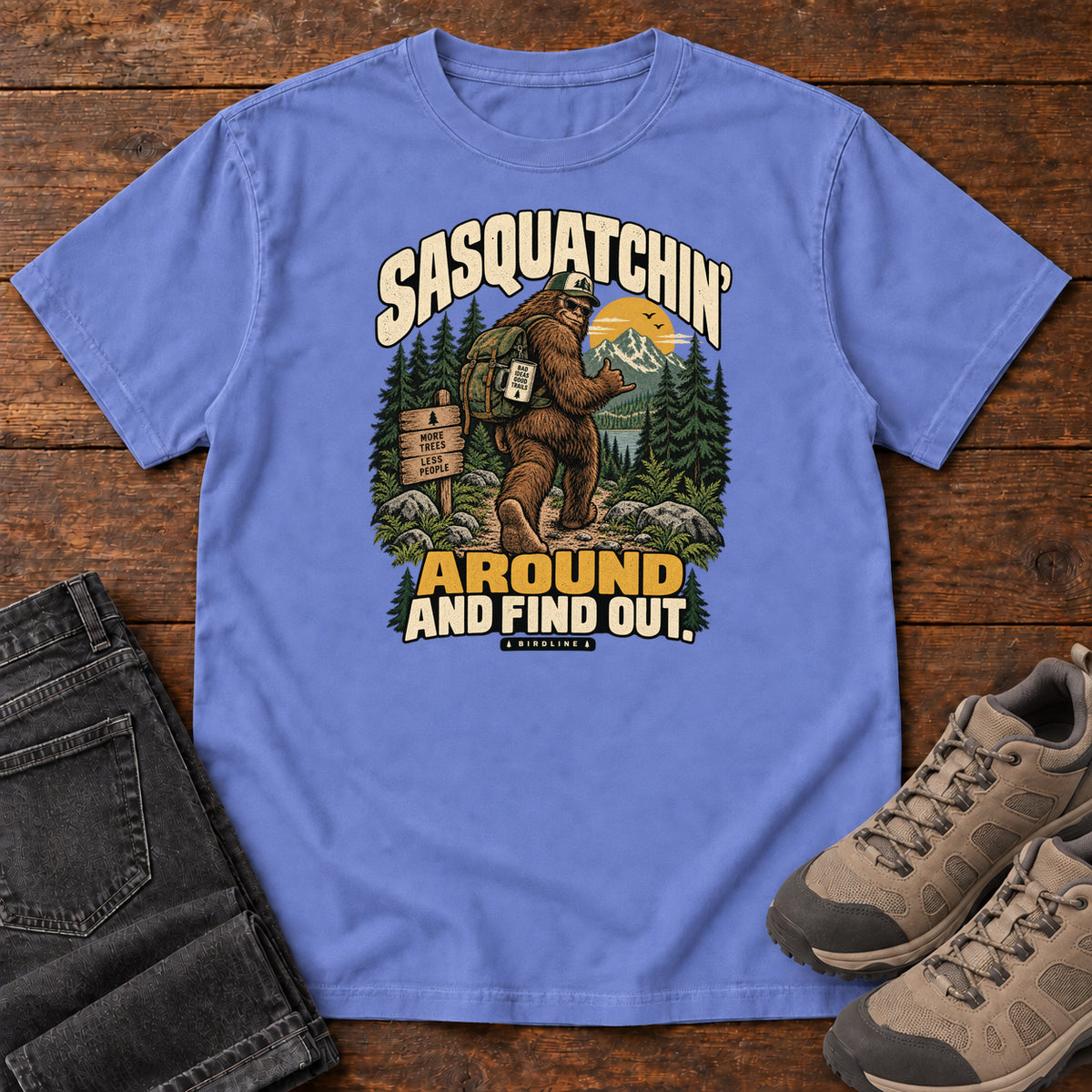 Flo Blue Sasquatchin Around and Find Out T-Shirt with a backpack-wearing Sasquatch on a forest trail with cream-and-gold lettering, front flat lay on wood.