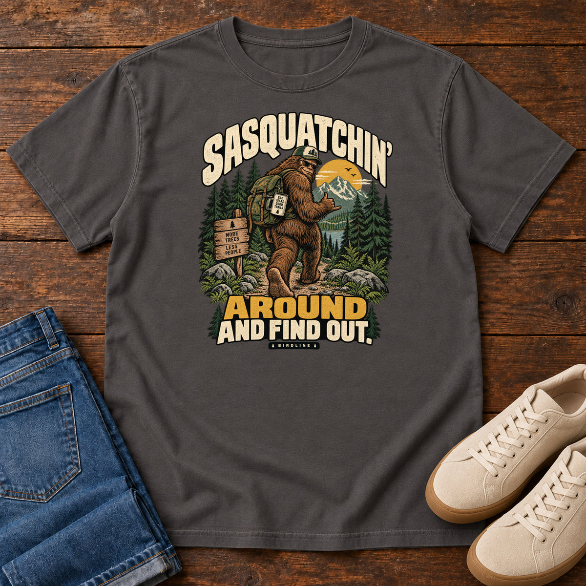 Graphite Sasquatchin Around and Find Out T-Shirt with a backpack-wearing Sasquatch on a forest trail with cream-and-gold lettering, front flat lay on wood.
