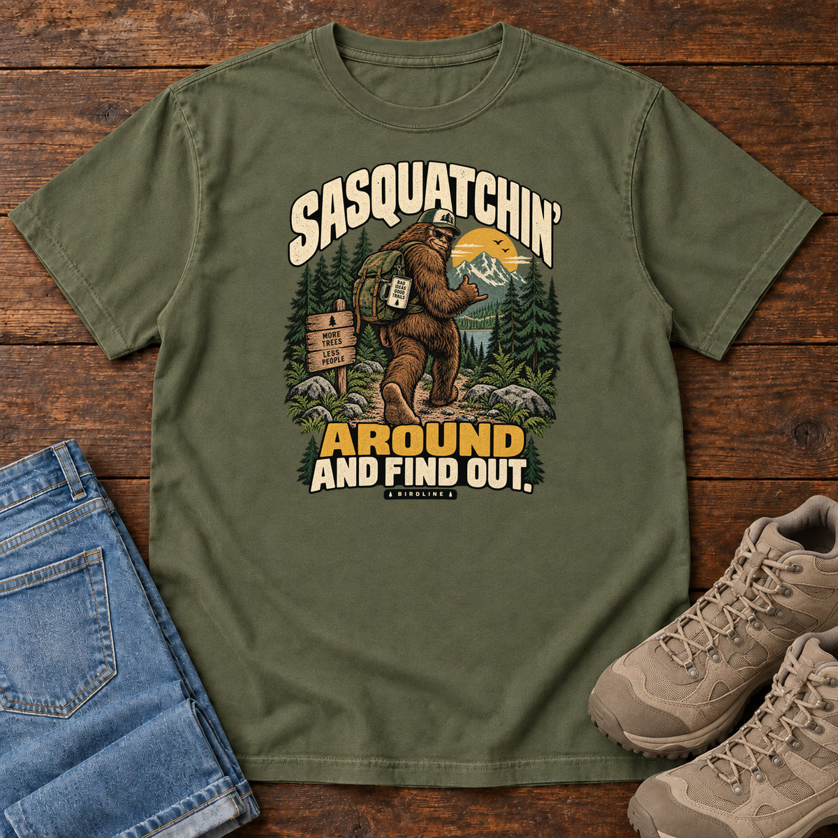 Hemp Sasquatchin Around and Find Out T-Shirt with a backpack-wearing Sasquatch on a forest trail with cream-and-gold lettering, front flat lay on wood.