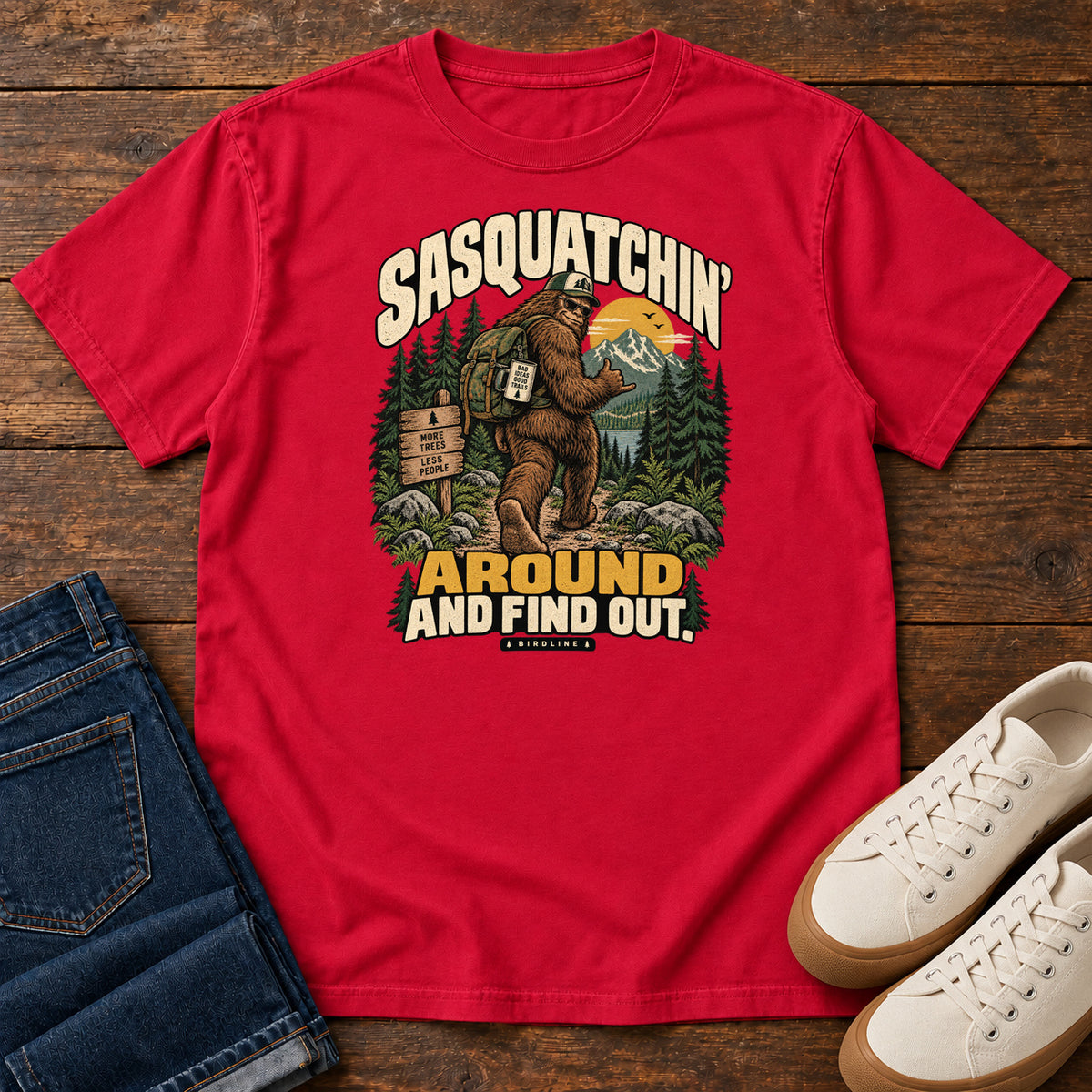 Red Sasquatchin Around and Find Out T-Shirt with a backpack-wearing Sasquatch on a forest trail with cream-and-gold lettering, front flat lay on wood.