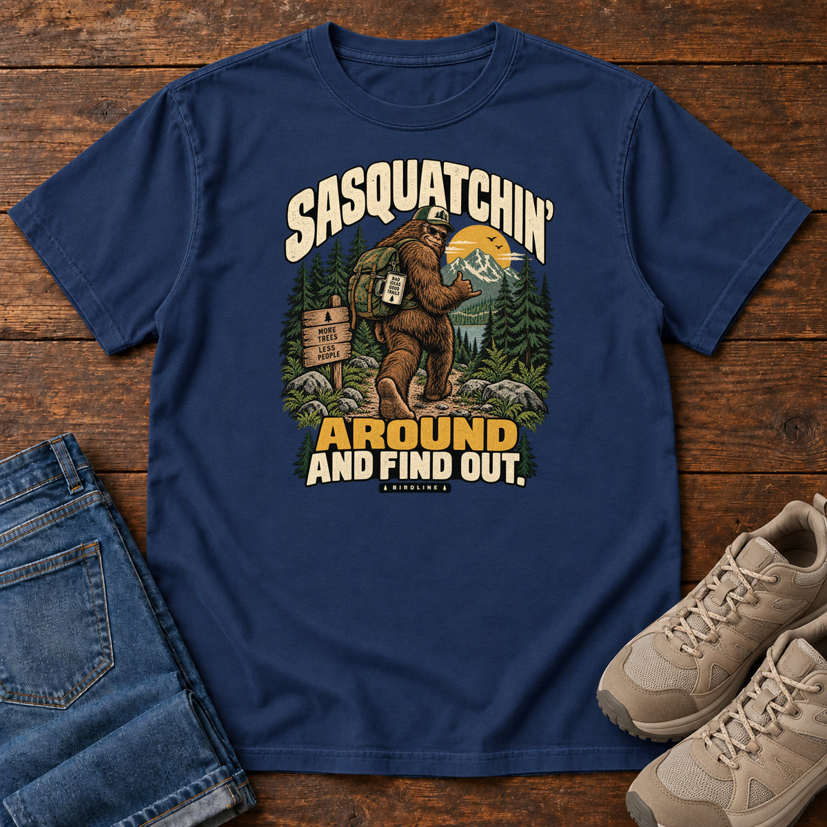 True Navy Sasquatchin Around and Find Out T-Shirt with a backpack-wearing Sasquatch on a forest trail with cream-and-gold lettering, front flat lay on wood.