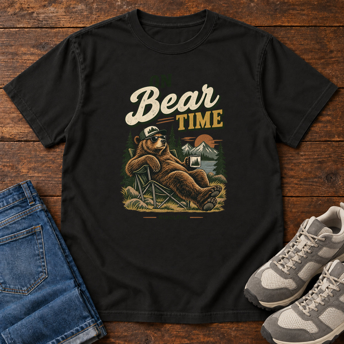 Black On Bear Time T-Shirt with a bear in a cap and sunglasses reclining in a camp chair with a mug; front flat lay on wood.