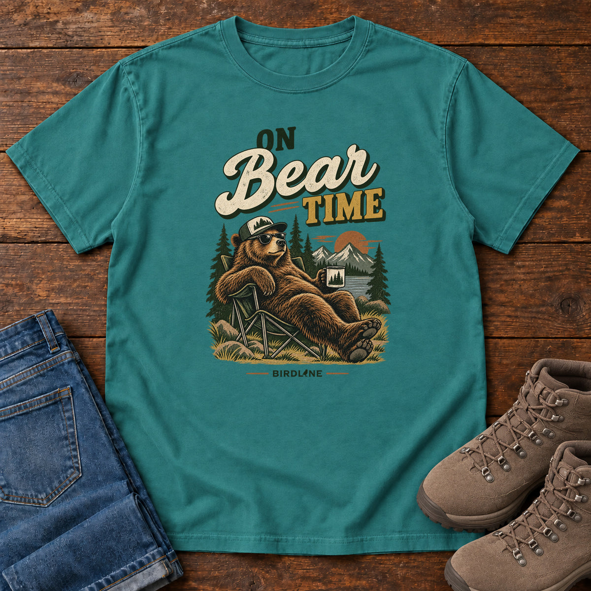 Emerald On Bear Time T-Shirt with a bear in a cap and sunglasses reclining in a camp chair with a mug; front flat lay on wood.