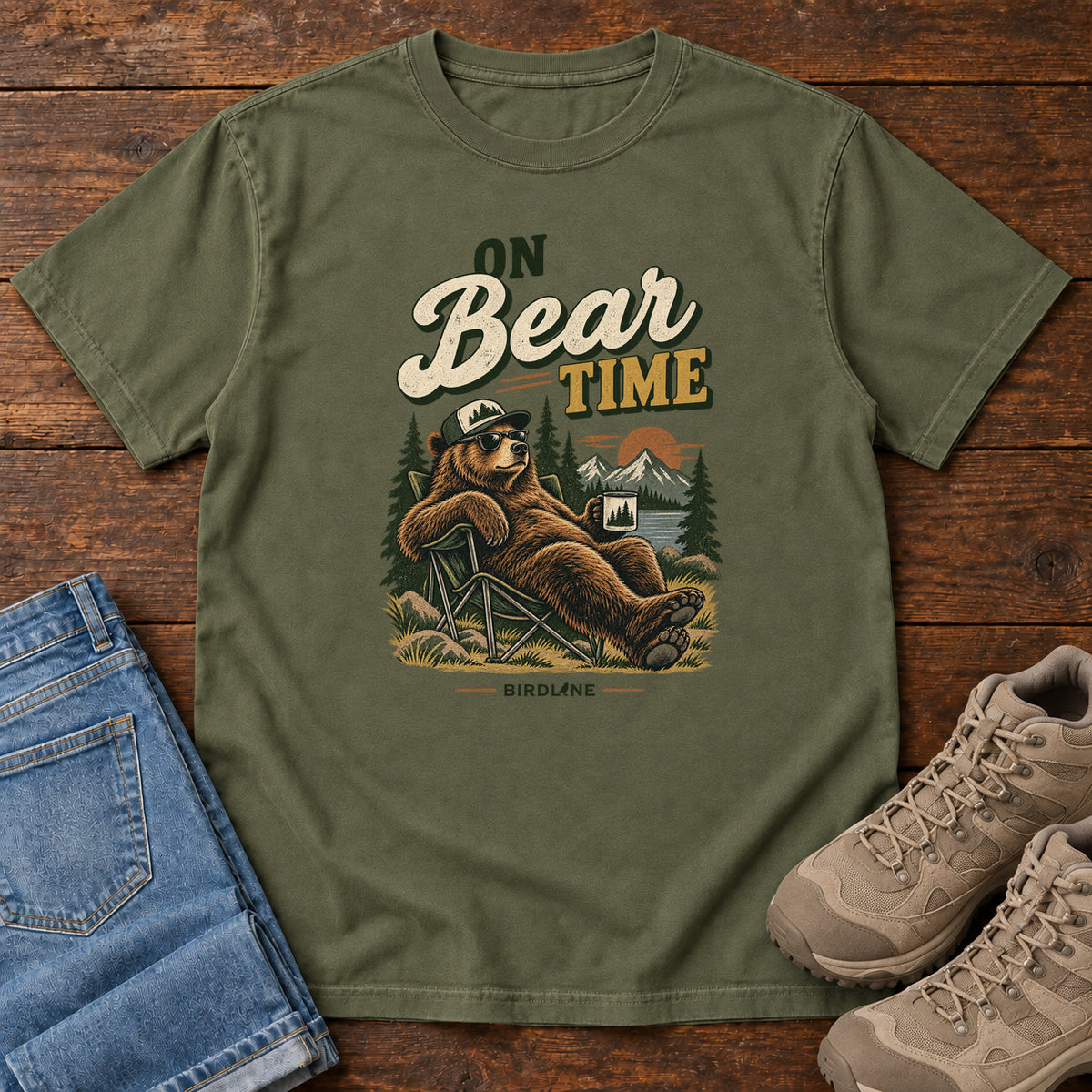 Hemp On Bear Time T-Shirt with a bear in a cap and sunglasses reclining in a camp chair with a mug; front flat lay on wood.
