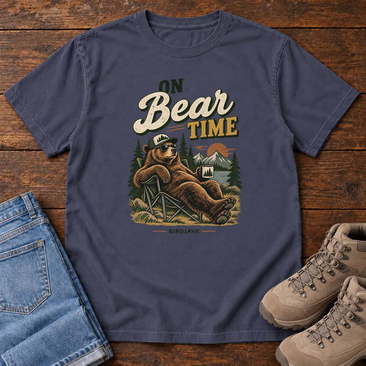 Navy On Bear Time T-Shirt with a bear in a cap and sunglasses reclining in a camp chair with a mug; front flat lay on wood.