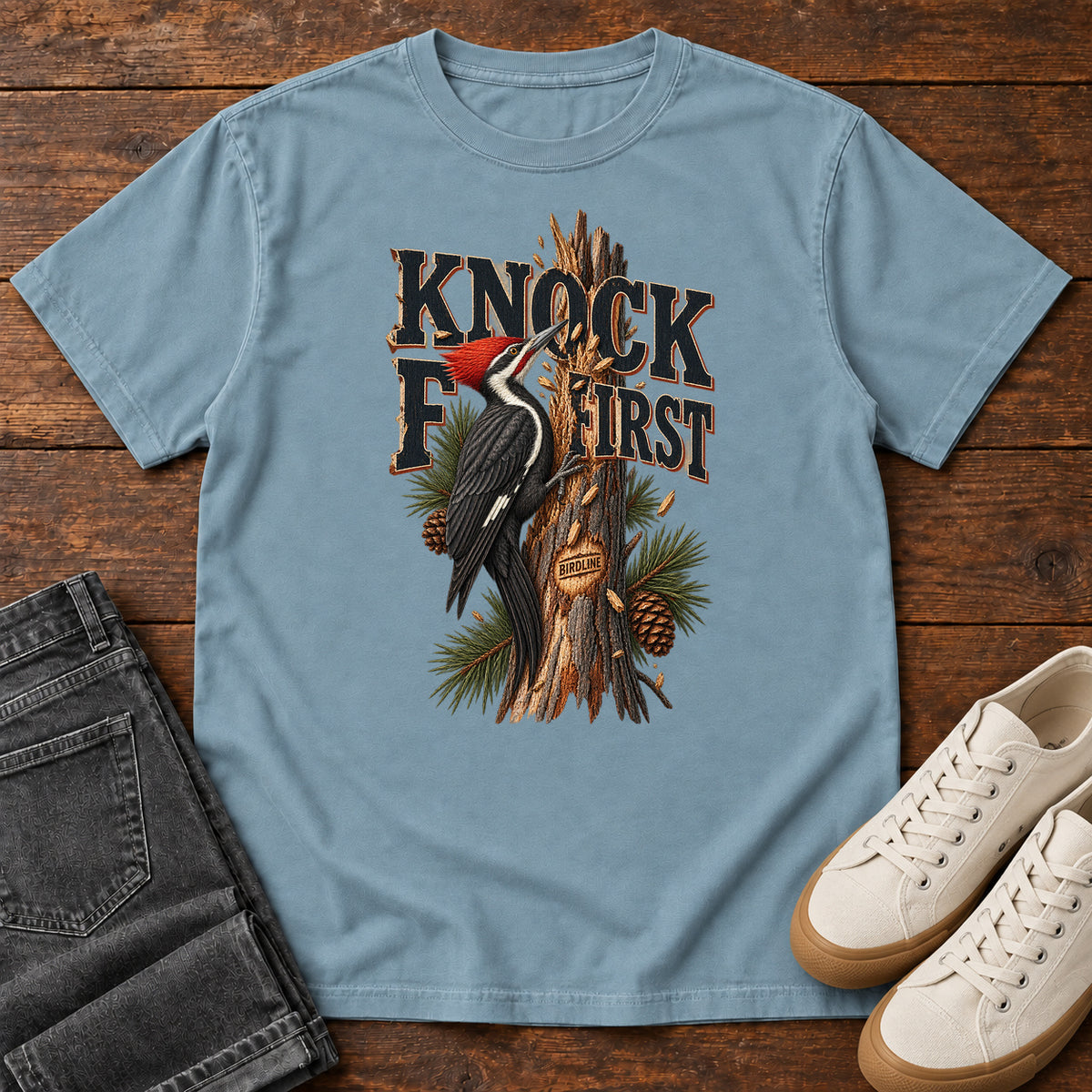 Ice Blue Knock First Woodpecker T-Shirt with a red-crested woodpecker on a pine trunk beneath Knock First lettering, front flat lay on wood.