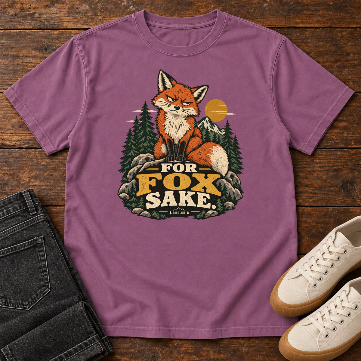 Berry For Fox Sake T-Shirt with an orange fox sitting on rocks against pines, mountains and a golden sun; front flat lay on wood.