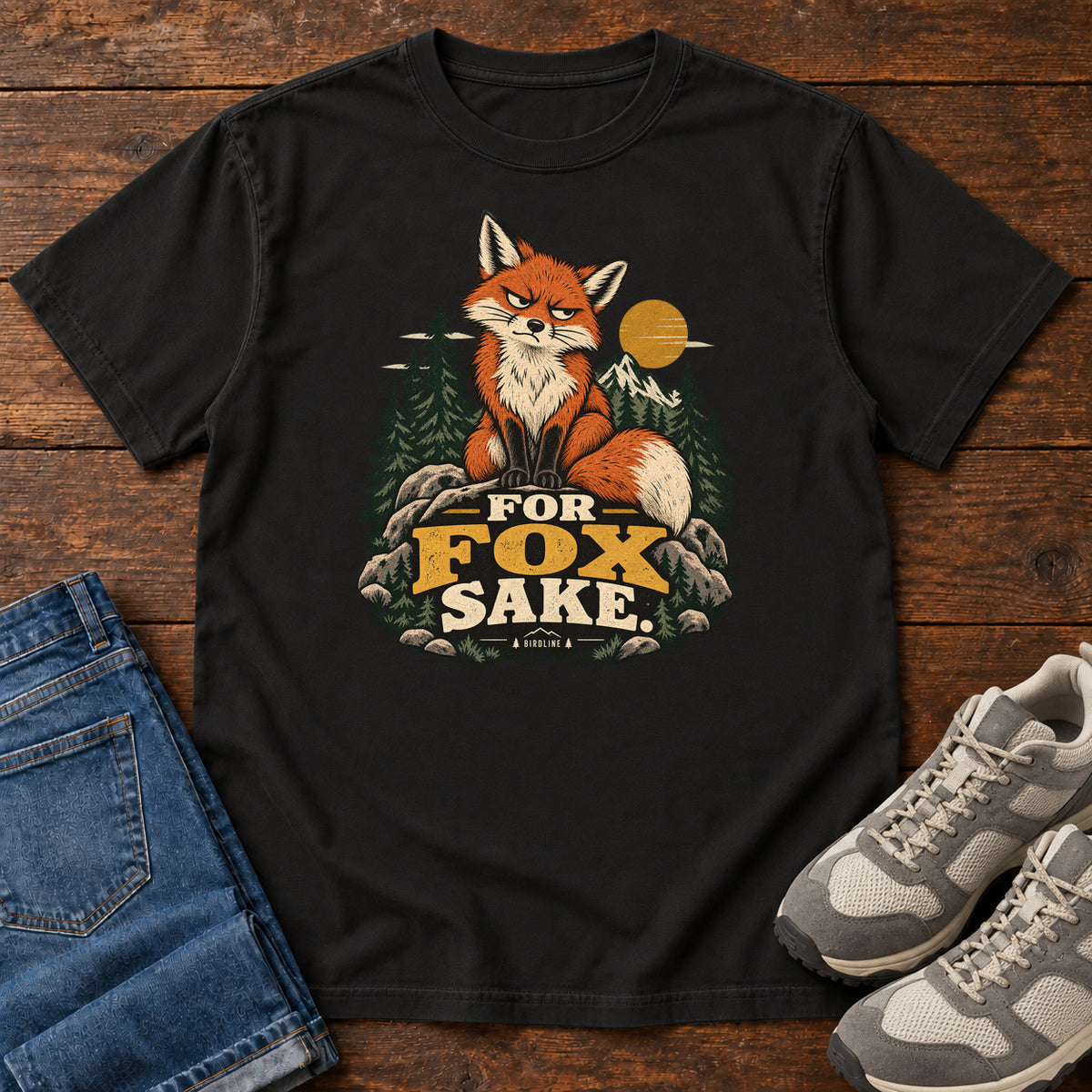 Black For Fox Sake T-Shirt with an orange fox sitting on rocks against pines, mountains and a golden sun; front flat lay on wood.