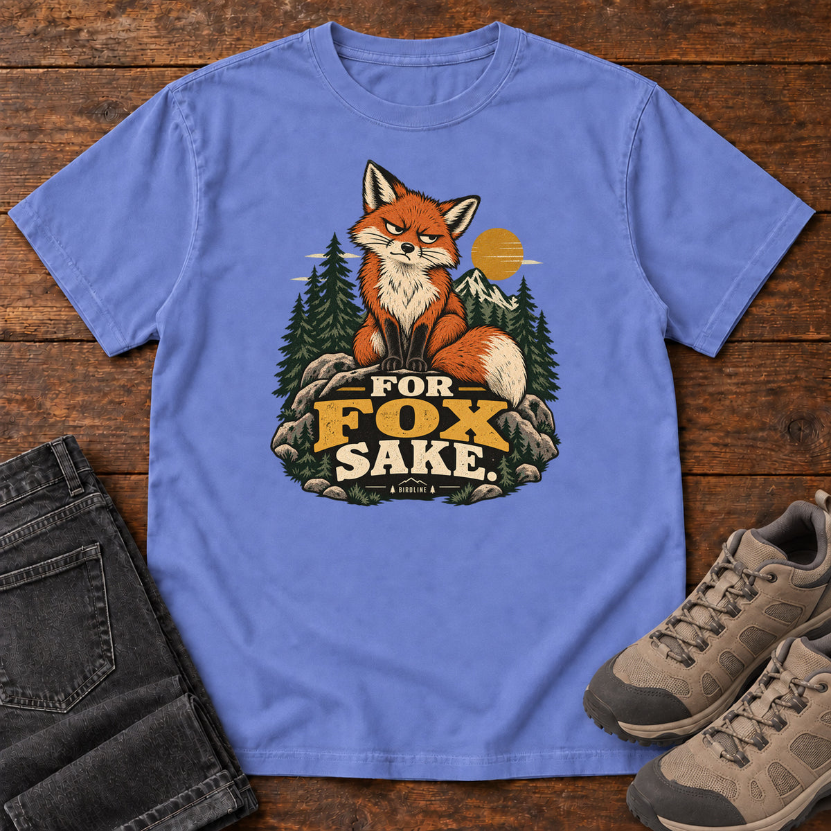 Flo Blue For Fox Sake T-Shirt with an orange fox sitting on rocks against pines, mountains and a golden sun; front flat lay on wood.