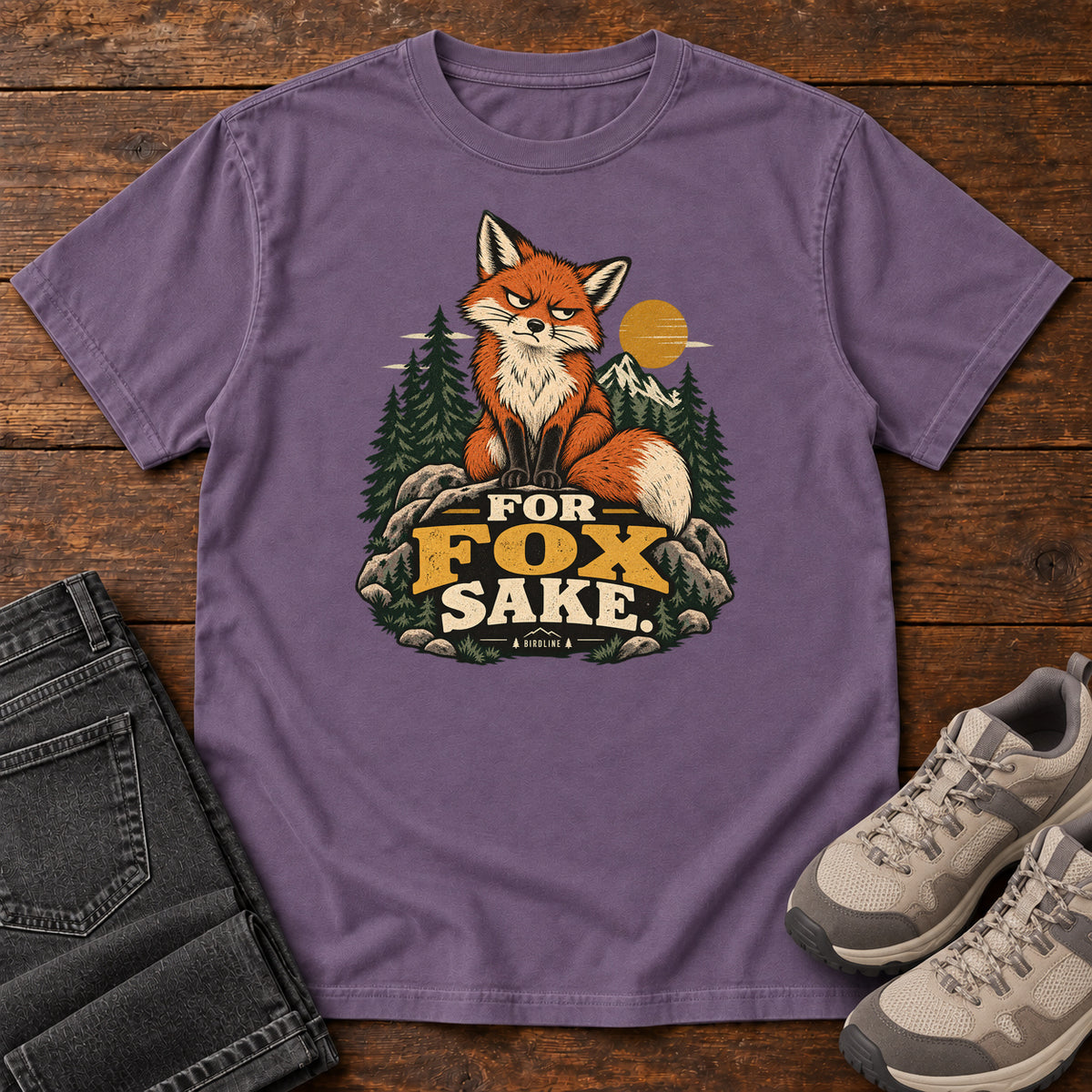 Grape For Fox Sake T-Shirt with an orange fox sitting on rocks against pines, mountains and a golden sun; front flat lay on wood.