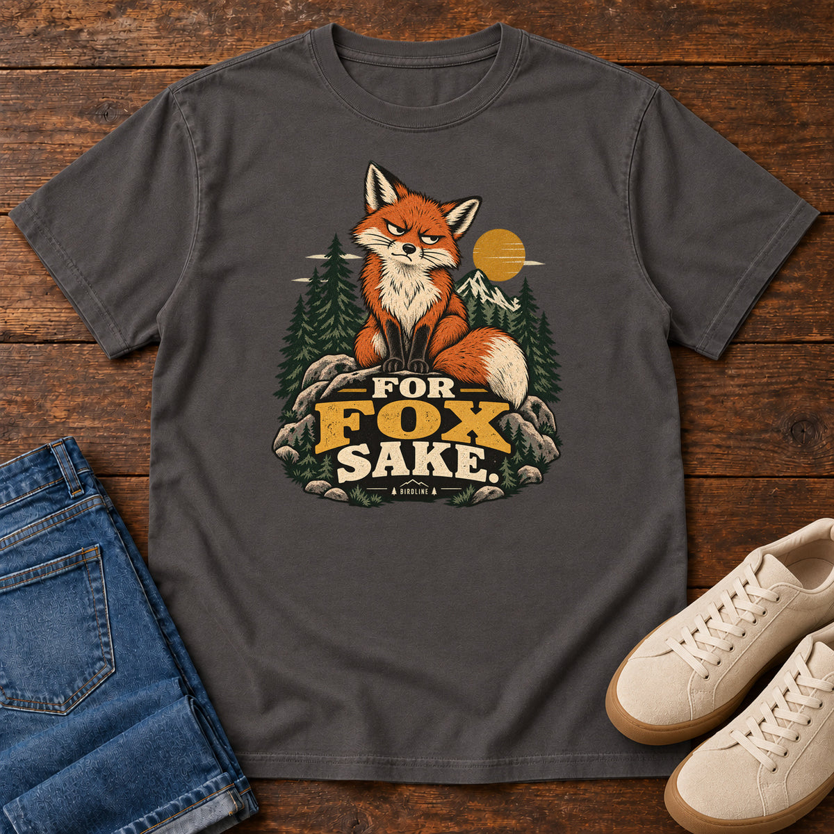 Graphite For Fox Sake T-Shirt with an orange fox sitting on rocks against pines, mountains and a golden sun; front flat lay on wood.