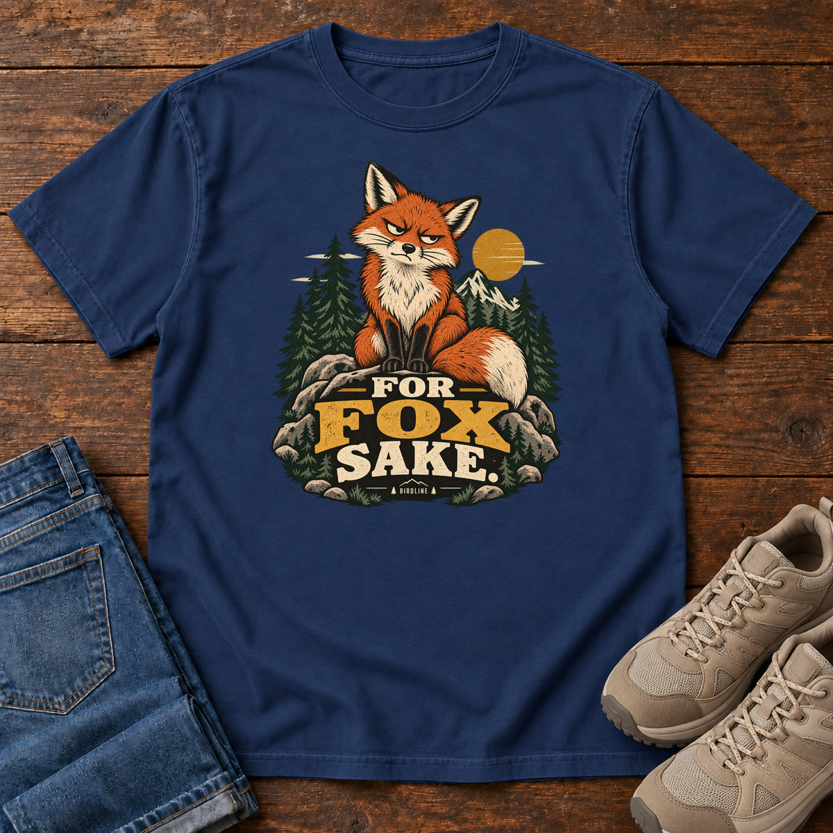 True Navy For Fox Sake T-Shirt with an orange fox sitting on rocks against pines, mountains and a golden sun; front flat lay on wood.