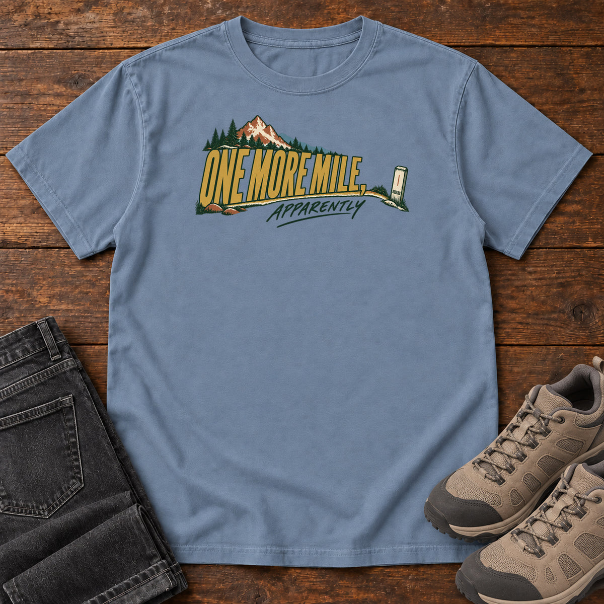 One More Mile Apparently T-Shirt in Blue Jean, front laid flat on wood, featuring gold slogan lettering with a mountain, pine trees and a small trail marker.