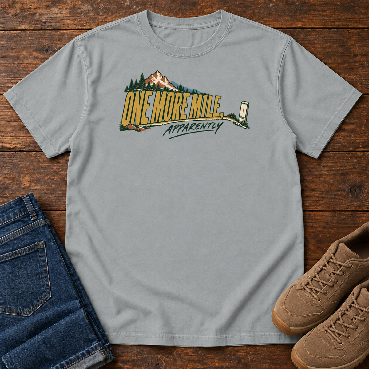 One More Mile Apparently T-Shirt in Granite, front laid flat on wood, featuring gold slogan lettering with a mountain, pine trees and a small trail marker.