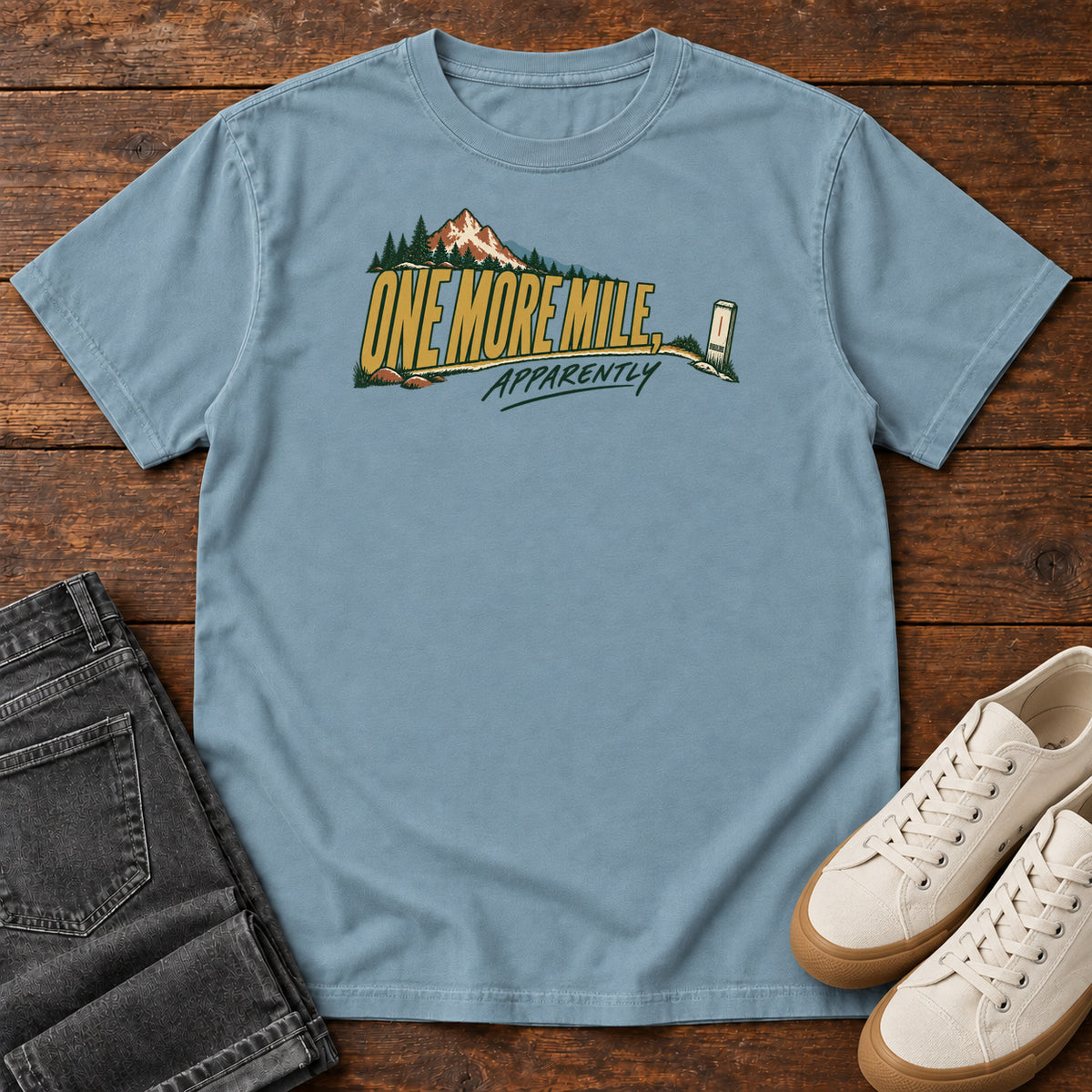 One More Mile Apparently T-Shirt in Ice Blue, front laid flat on wood, featuring gold slogan lettering with a mountain, pine trees and a small trail marker.