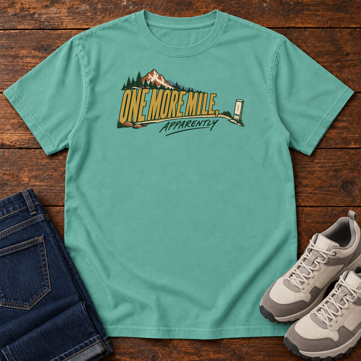One More Mile Apparently T-Shirt in Seafoam, front laid flat on wood, featuring gold slogan lettering with a mountain, pine trees and a small trail marker.