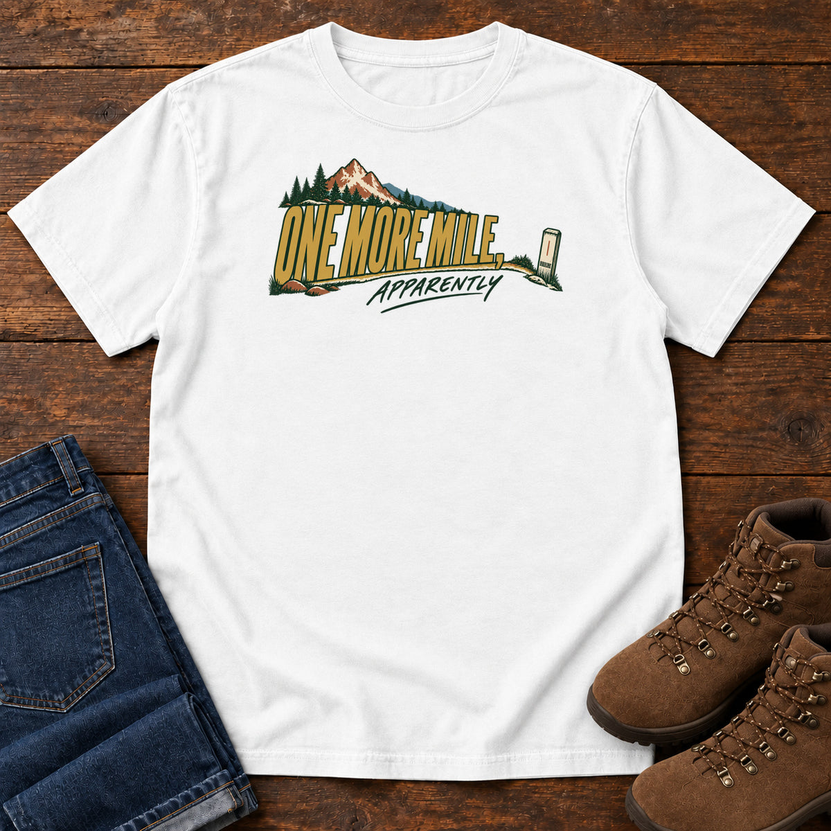 One More Mile Apparently T-Shirt in White, front laid flat on wood, featuring gold slogan lettering with a mountain, pine trees and a small trail marker.