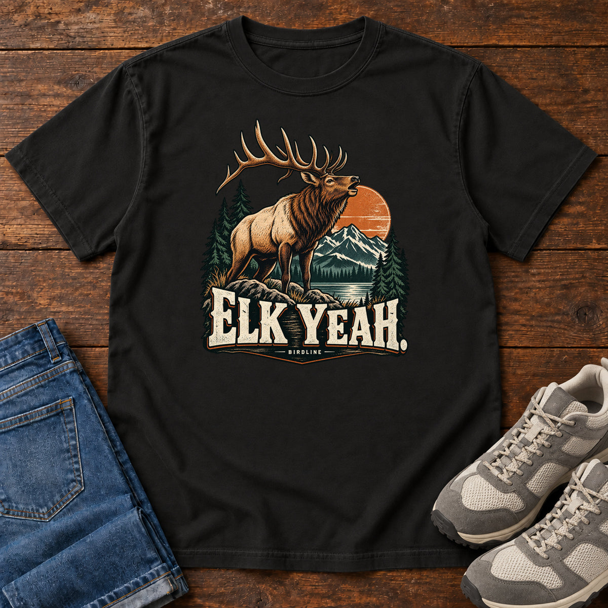 Elk Yeah T-Shirt in Black, front laid flat on wood, featuring an antlered elk before mountains, pine trees and an orange sun, above cream lettering.