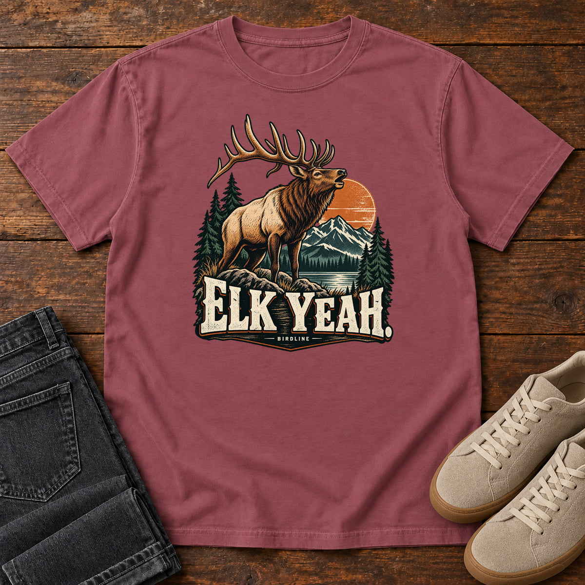 Elk Yeah T-Shirt in Brick, front laid flat on wood, featuring an antlered elk before mountains, pine trees and an orange sun, above cream lettering.