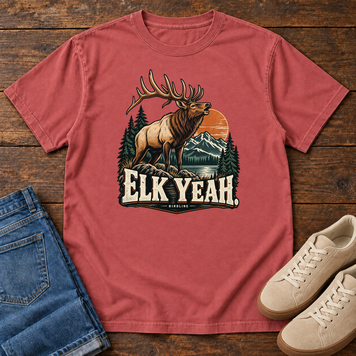 Elk Yeah T-Shirt in Crimson, front laid flat on wood, featuring an antlered elk before mountains, pine trees and an orange sun, above cream lettering.