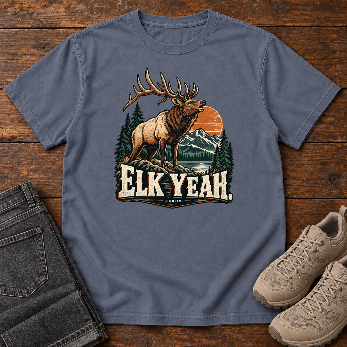 Elk Yeah T-Shirt in Denim, front laid flat on wood, featuring an antlered elk before mountains, pine trees and an orange sun, above cream lettering.