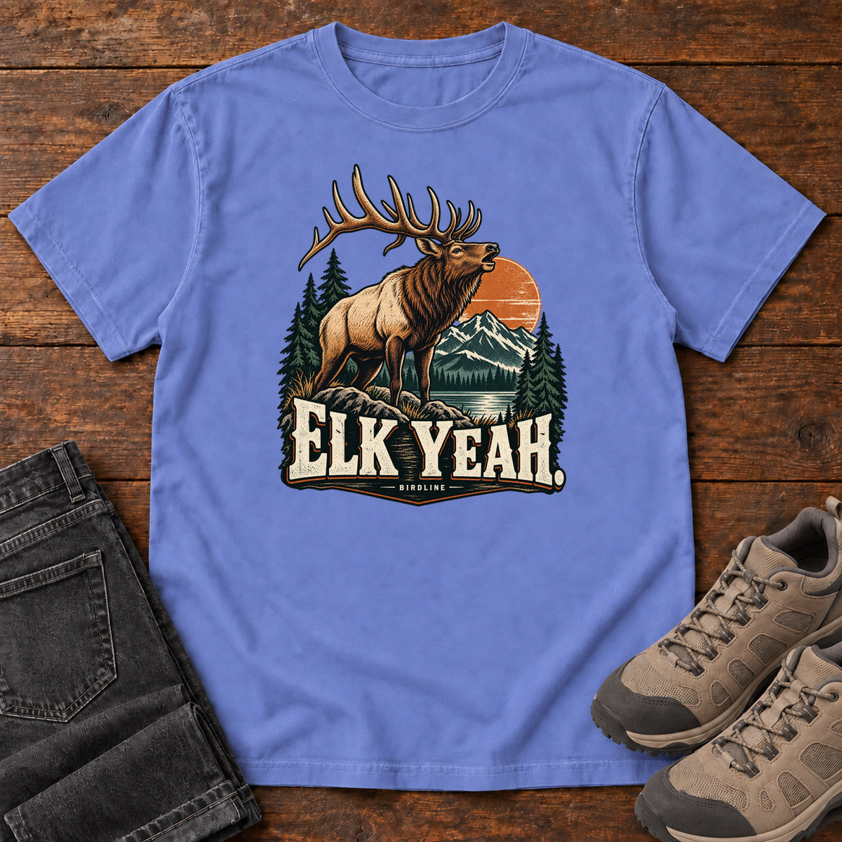 Elk Yeah T-Shirt in Flo Blue, front laid flat on wood, featuring an antlered elk before mountains, pine trees and an orange sun, above cream lettering.