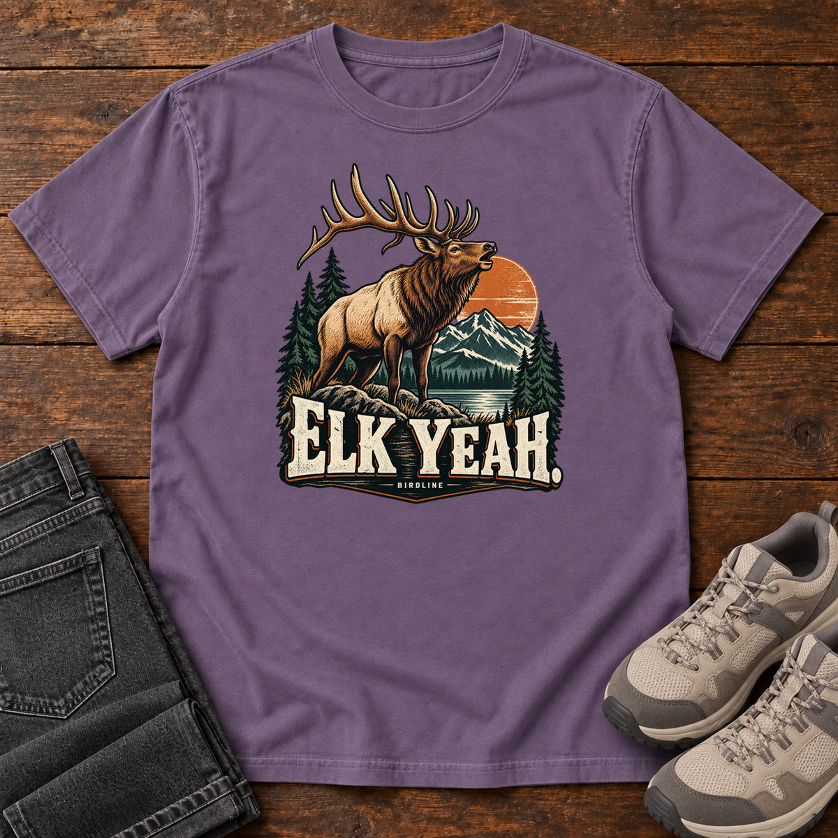 Elk Yeah T-Shirt in Grape, front laid flat on wood, featuring an antlered elk before mountains, pine trees and an orange sun, above cream lettering.