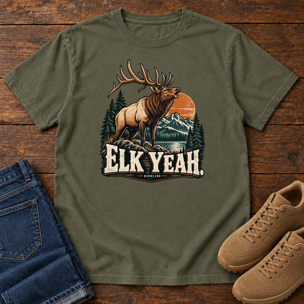 Elk Yeah T-Shirt in Sage, front laid flat on wood, featuring an antlered elk before mountains, pine trees and an orange sun, above cream lettering.