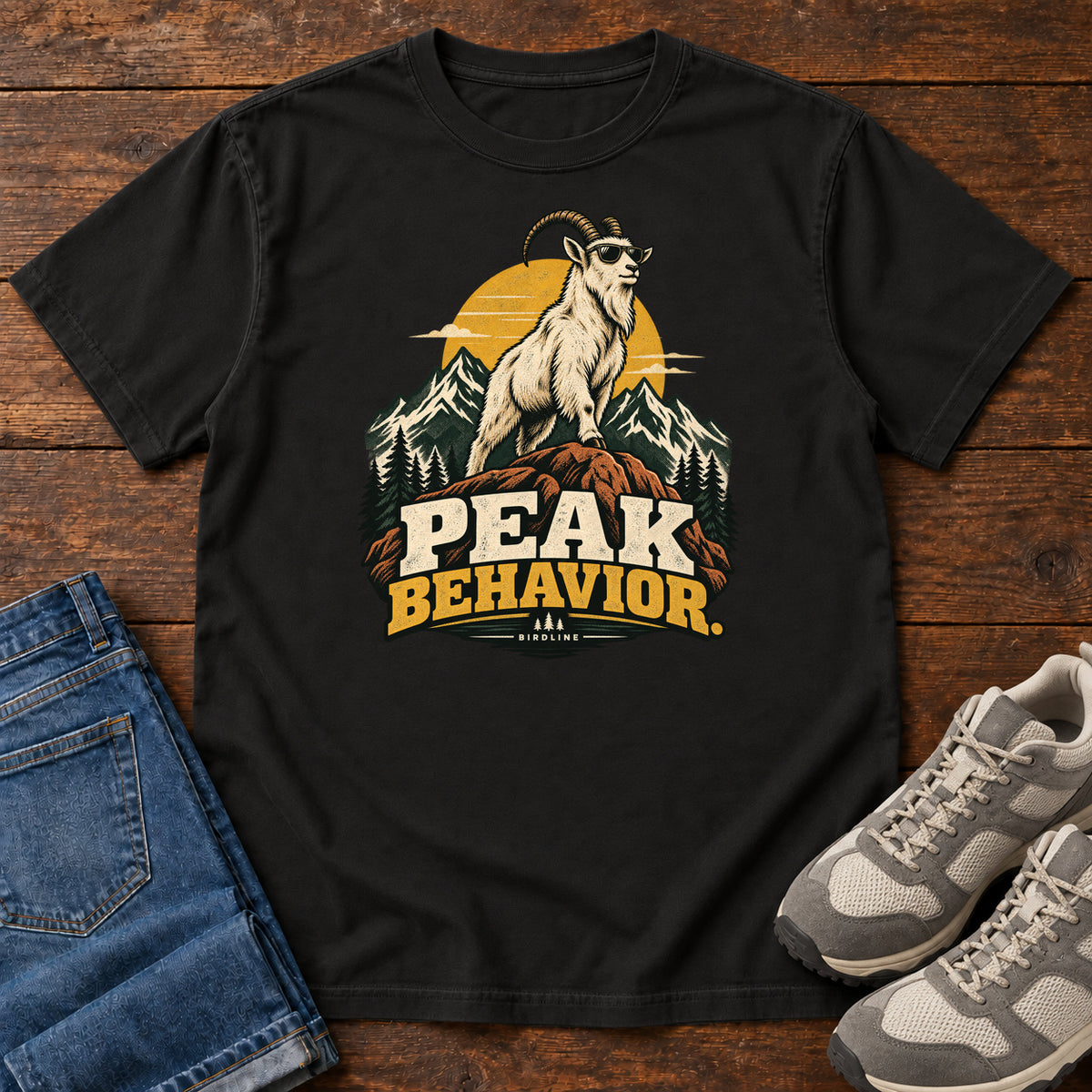 Peak Behavior T-Shirt in Black, front laid flat on wood, featuring a horned white goat wearing sunglasses on a rocky peak, with mountains, a gold sun and bold lettering.