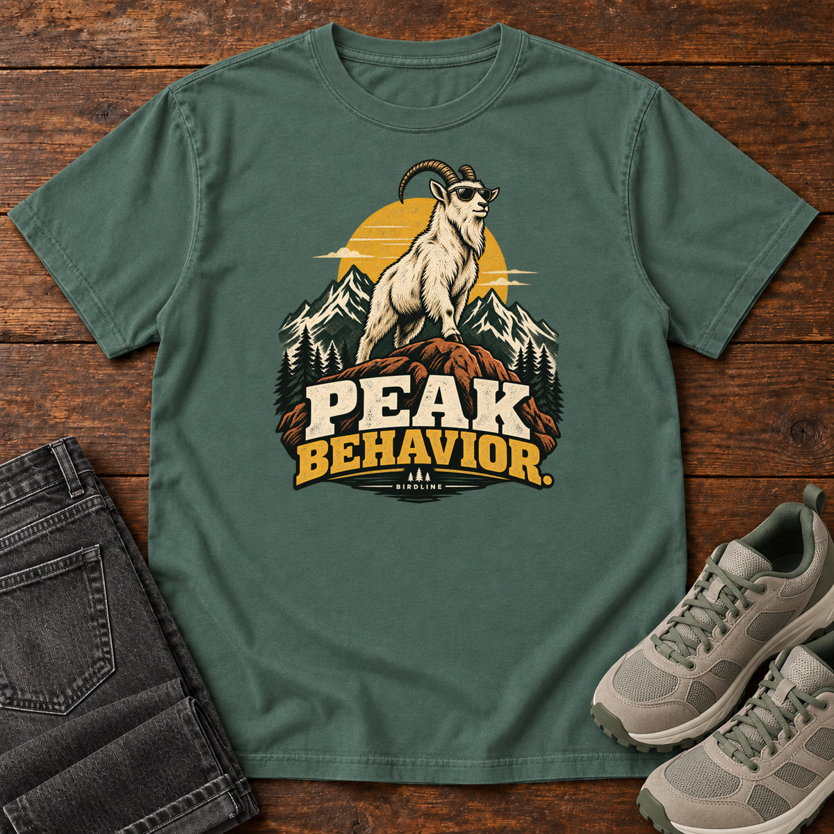 Peak Behavior T-Shirt in Blue Spruce, front laid flat on wood, featuring a horned white goat wearing sunglasses on a rocky peak, with mountains, a gold sun and bold lettering.
