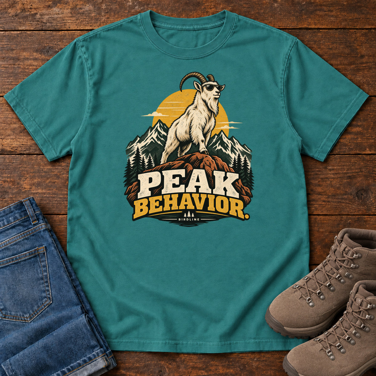 Peak Behavior T-Shirt in Emerald, front laid flat on wood, featuring a horned white goat wearing sunglasses on a rocky peak, with mountains, a gold sun and bold lettering.