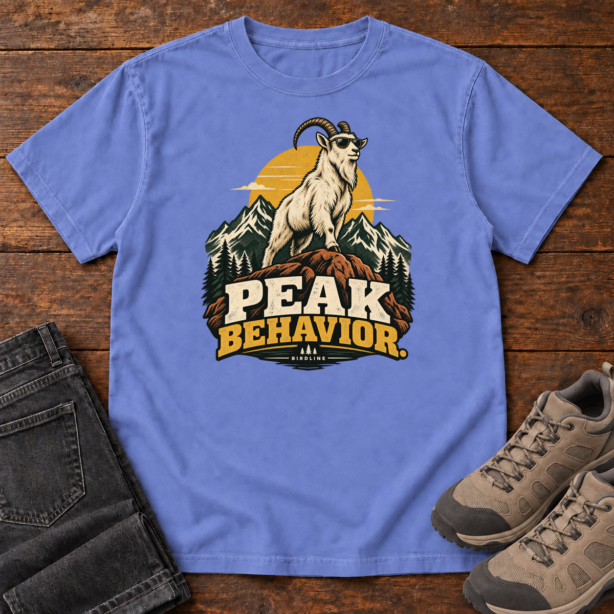 Peak Behavior T-Shirt in Flo Blue, front laid flat on wood, featuring a horned white goat wearing sunglasses on a rocky peak, with mountains, a gold sun and bold lettering.