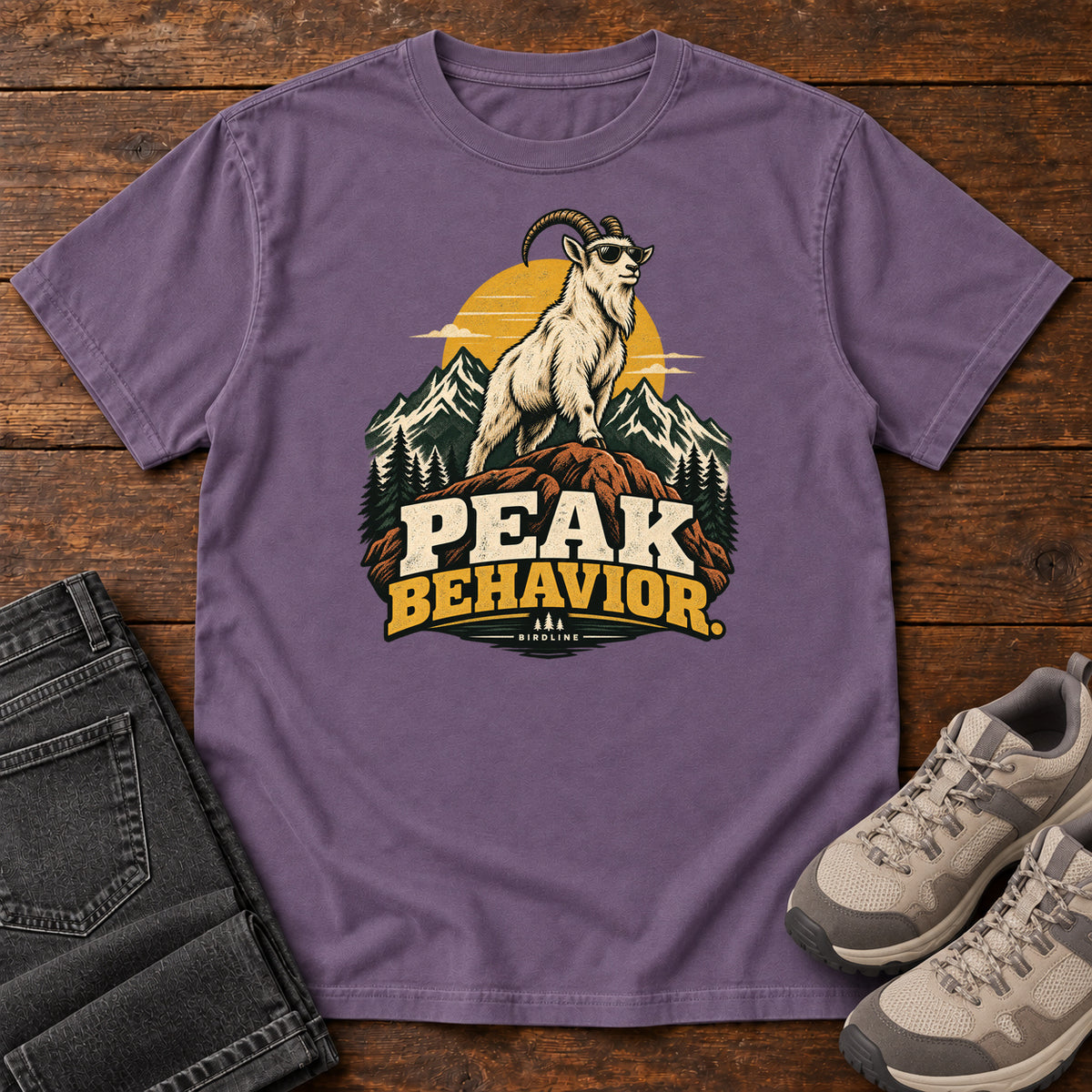 Peak Behavior T-Shirt in Grape, front laid flat on wood, featuring a horned white goat wearing sunglasses on a rocky peak, with mountains, a gold sun and bold lettering.