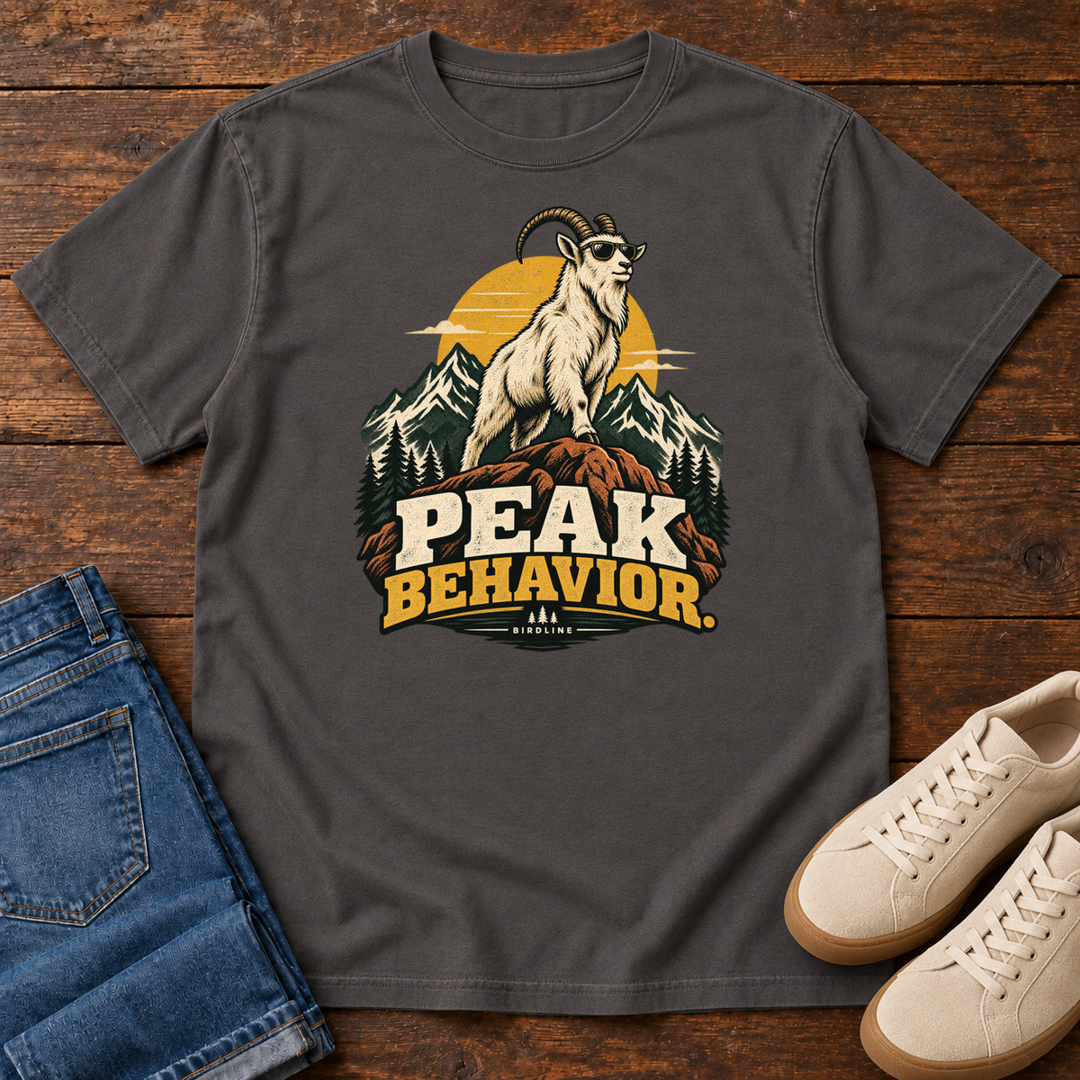 Peak Behavior T-Shirt in Graphite, front laid flat on wood, featuring a horned white goat wearing sunglasses on a rocky peak, with mountains, a gold sun and bold lettering.
