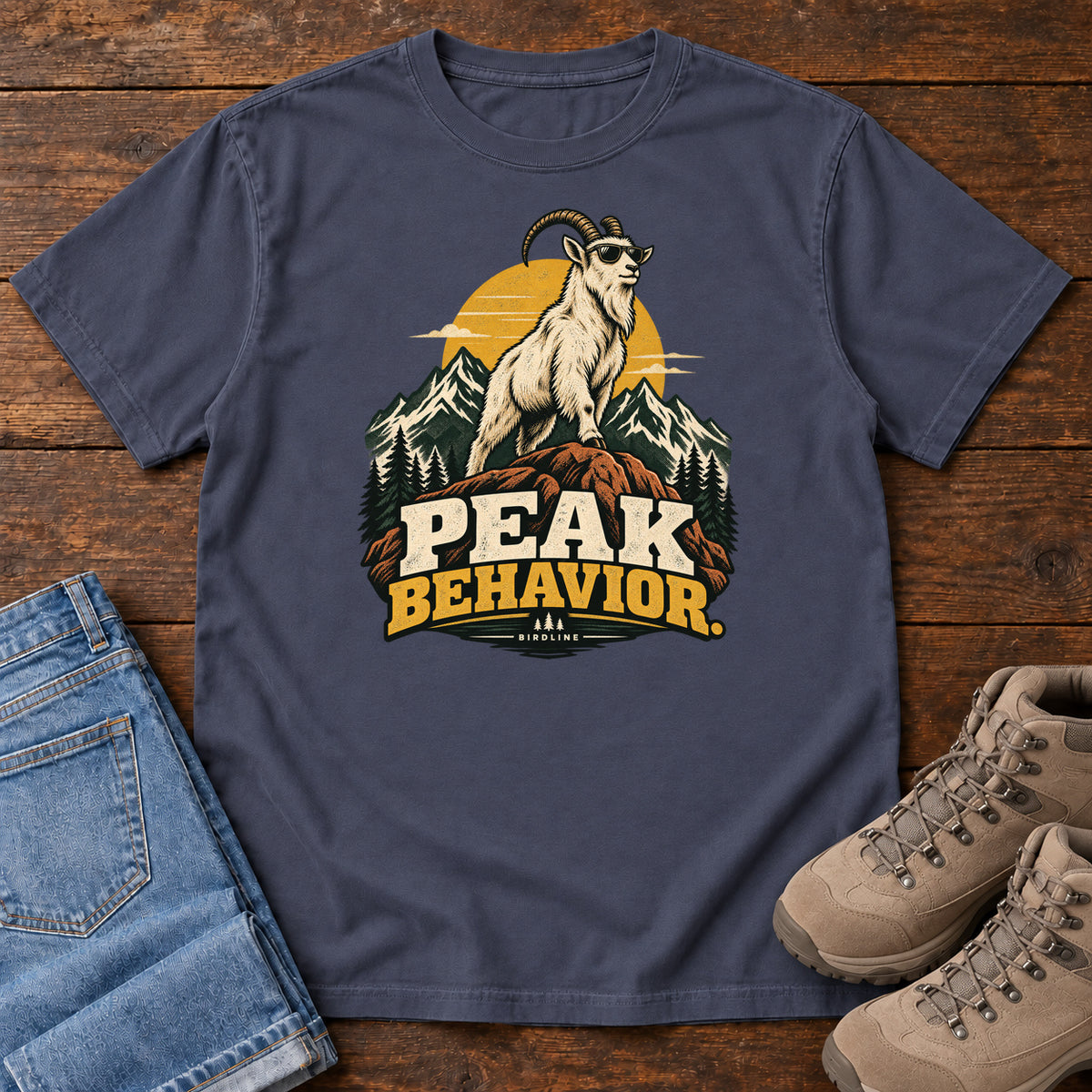 Peak Behavior T-Shirt in Navy, front laid flat on wood, featuring a horned white goat wearing sunglasses on a rocky peak, with mountains, a gold sun and bold lettering.